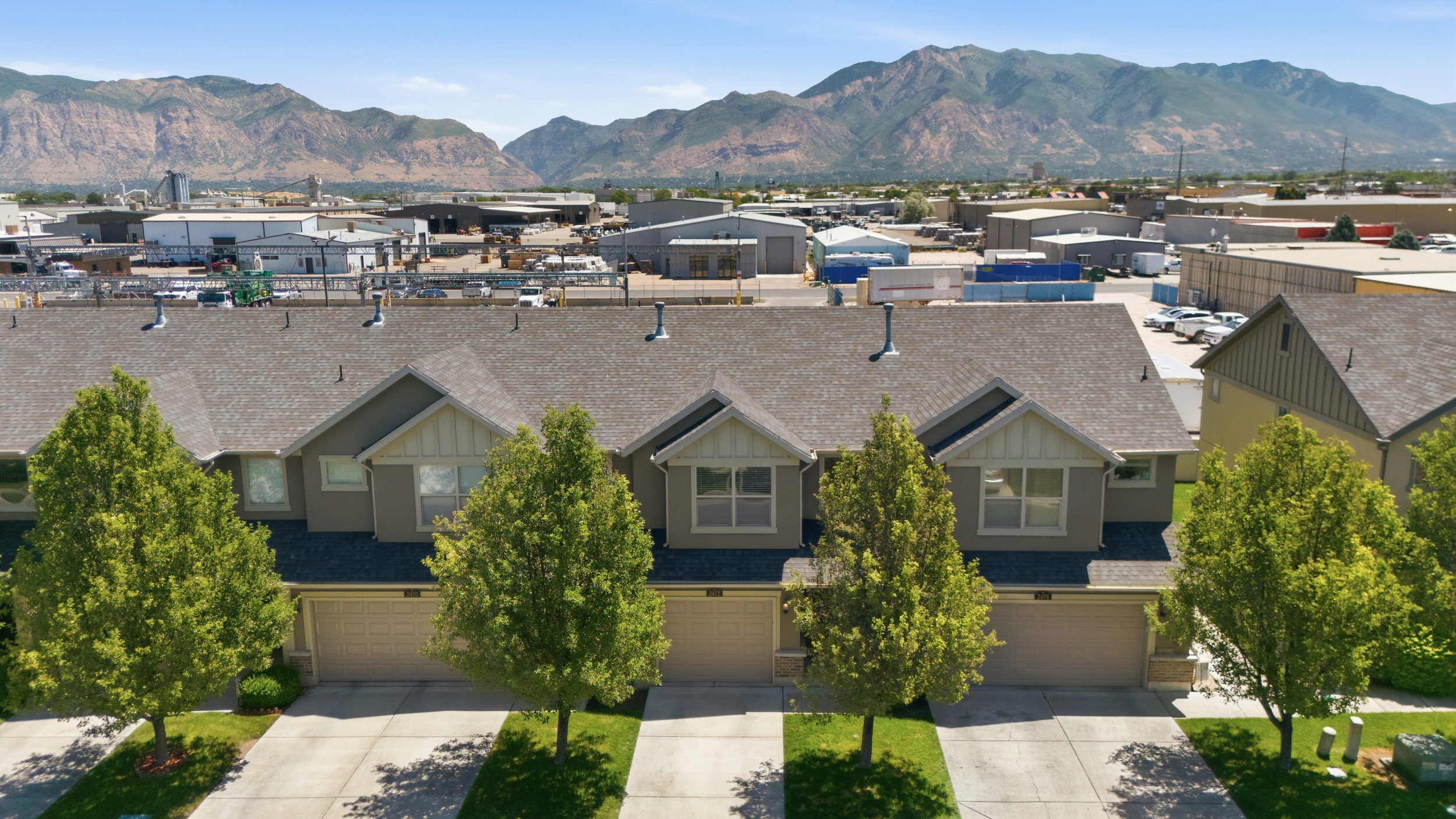 Modern townhome exterior featuring an attached garage, landscaped front yard with mature trees, and a gray shingle roof