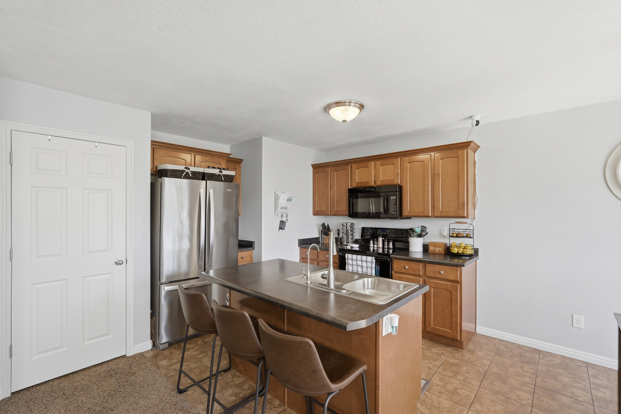 Kitchen with wood cabinetry, stainless steel appliances, a black countertop, and an island featuring a double-basin sink