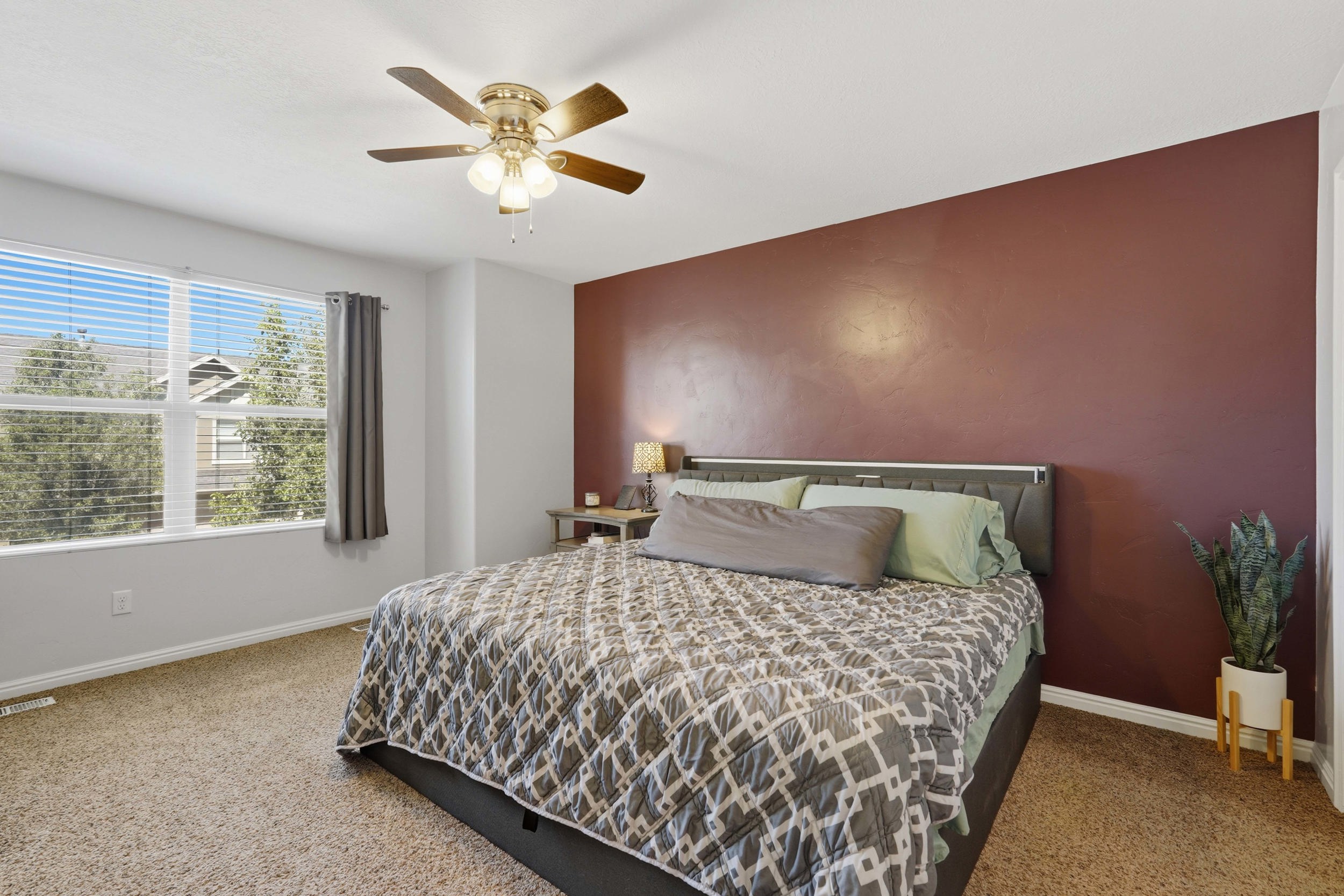 Carpeted interior space featuring a large window with horizontal blinds, a ceiling fan with integrated lighting, and a prominent accent wall