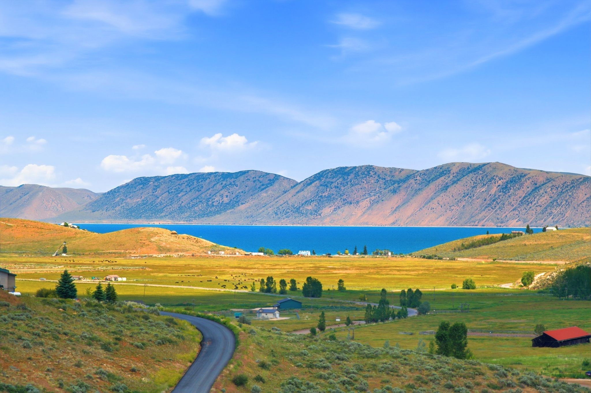 Expansive landscape featuring a winding asphalt road, verdant hillsides, distant blue lake, and mountain range backdrop
