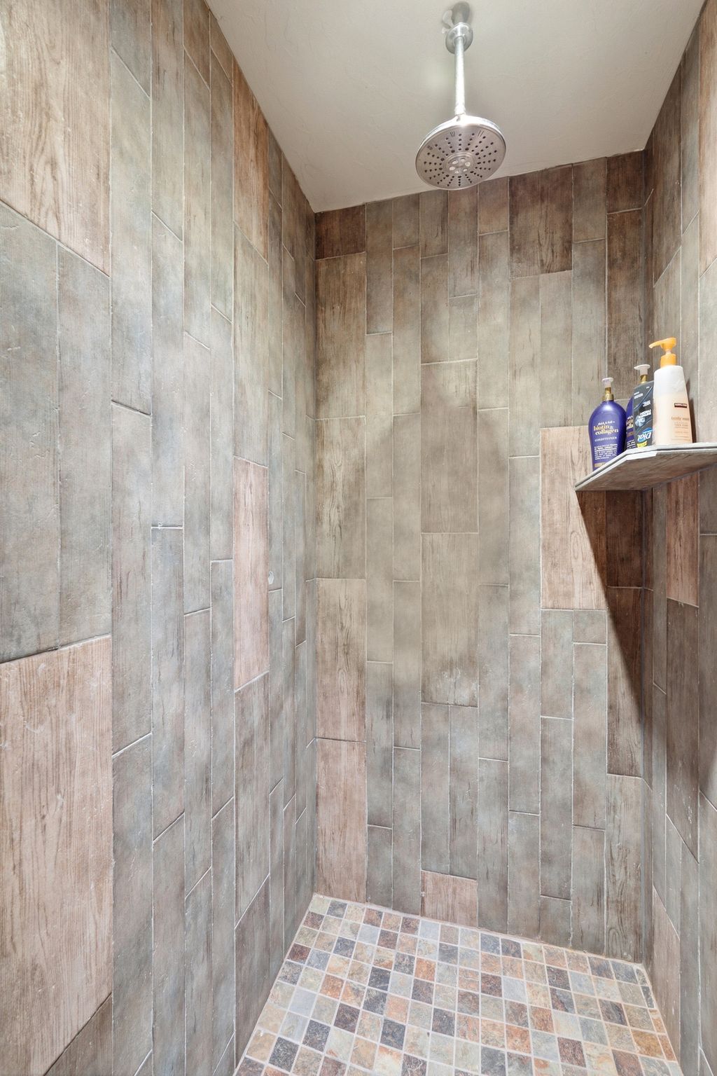Walk-in shower featuring full-height tiled walls, a ceiling-mounted rainfall showerhead, a built-in tiled shelf, and a mosaic-tiled floor