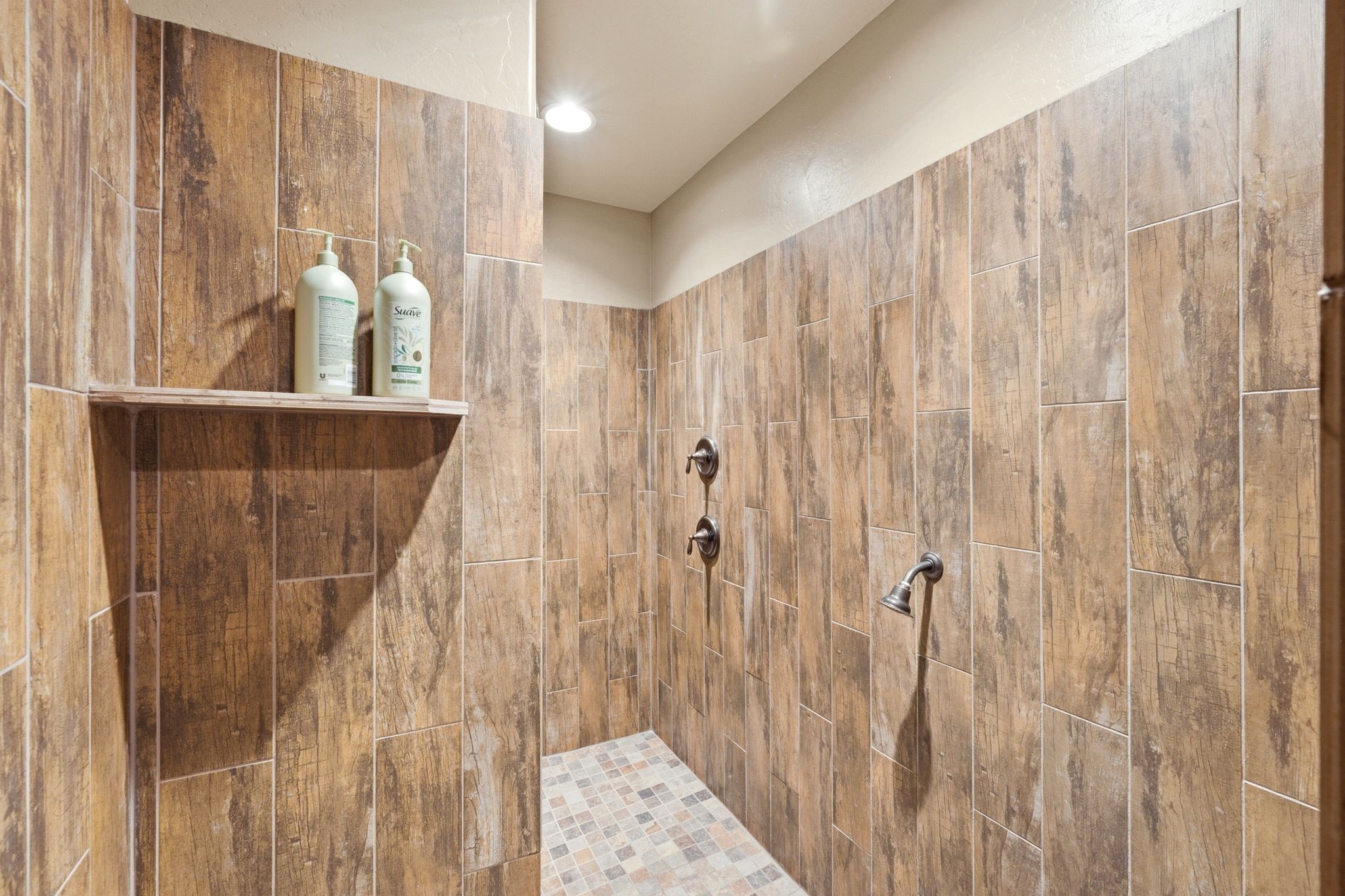 Spacious walk-in shower featuring wood-finish tile walls, dual shower heads, and a mosaic tile floor