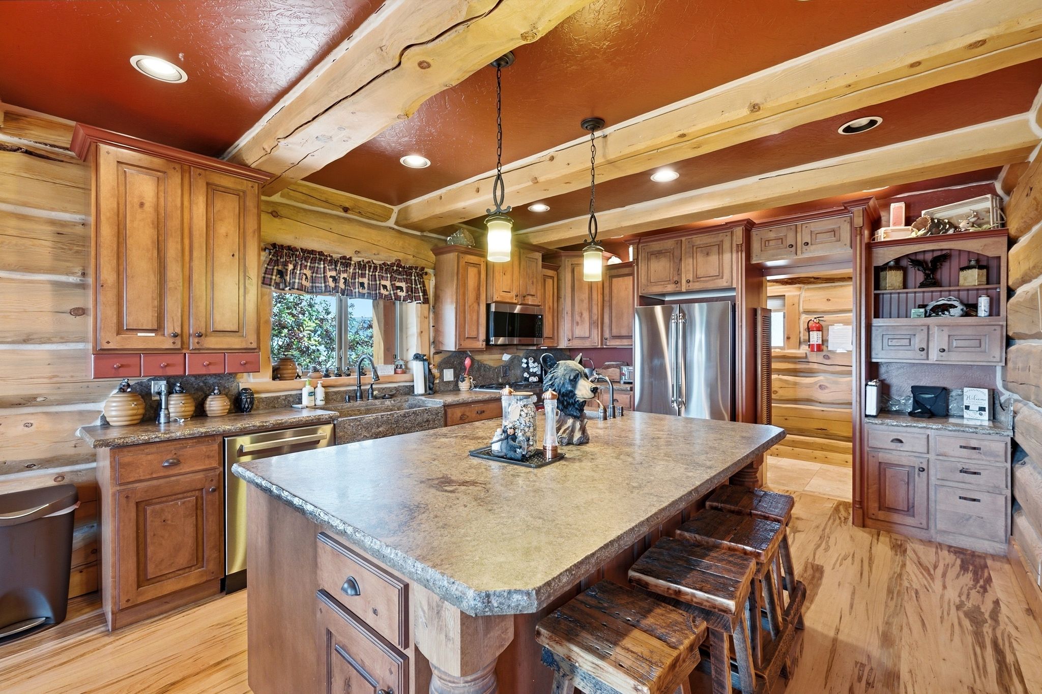 Spacious kitchen featuring a large central island with stone-finish countertop, stainless steel appliances, wood-finish flooring, and exposed log beam ceilings