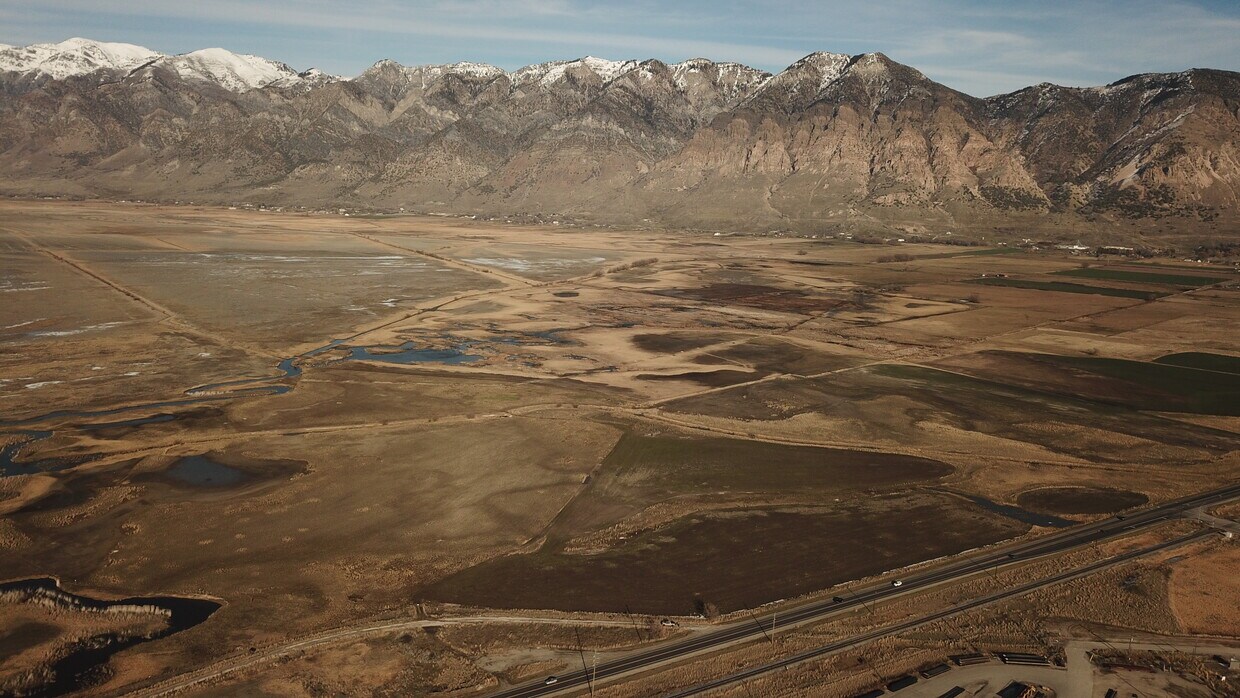 Expansive high desert landscape featuring a prominent mountain range with snow-capped peaks, and a highway traversing the foreground