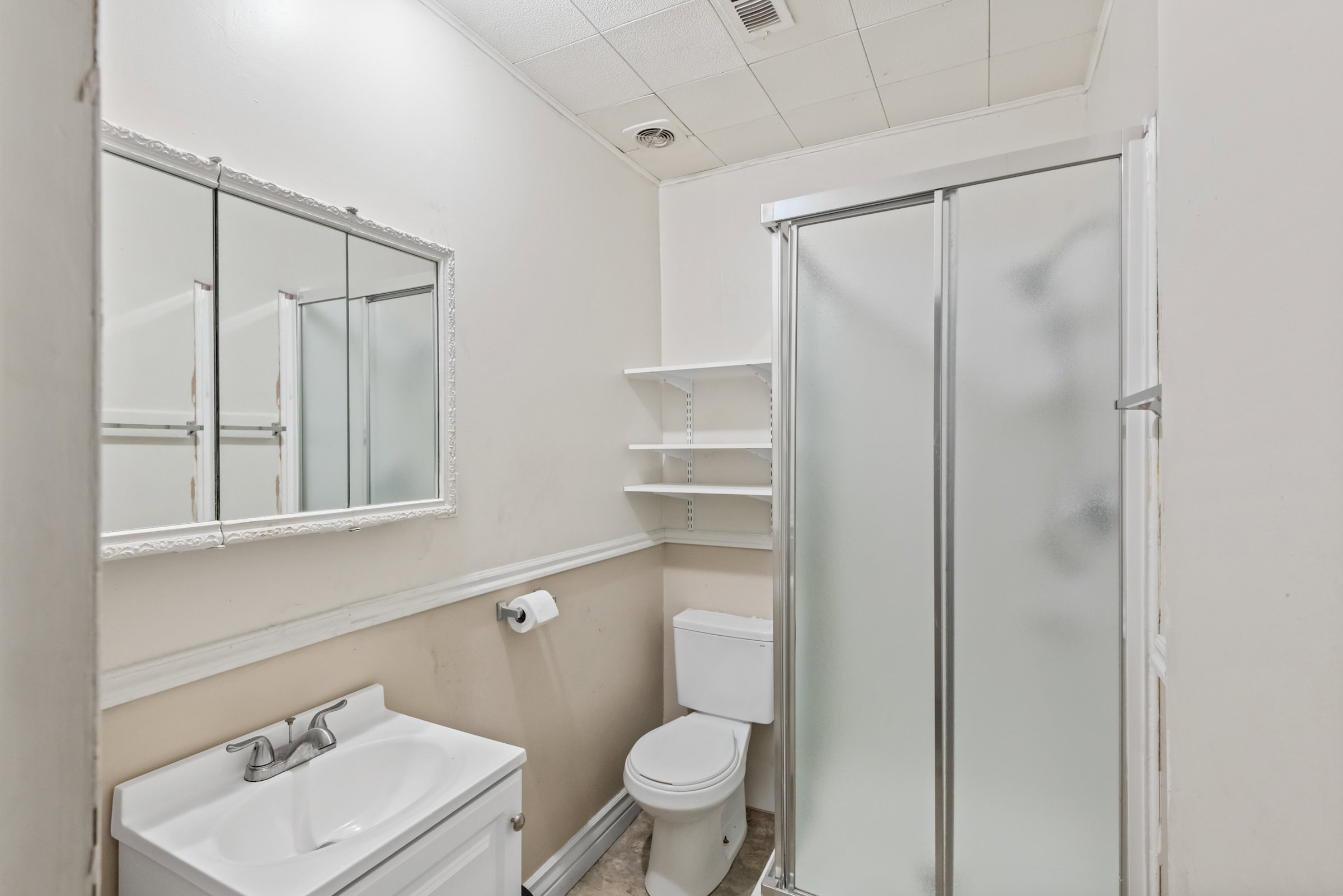 Bathroom featuring a white vanity with integrated sink, a framed medicine cabinet, a toilet, a shower with frosted glass enclosure, and wall-mounted shelving