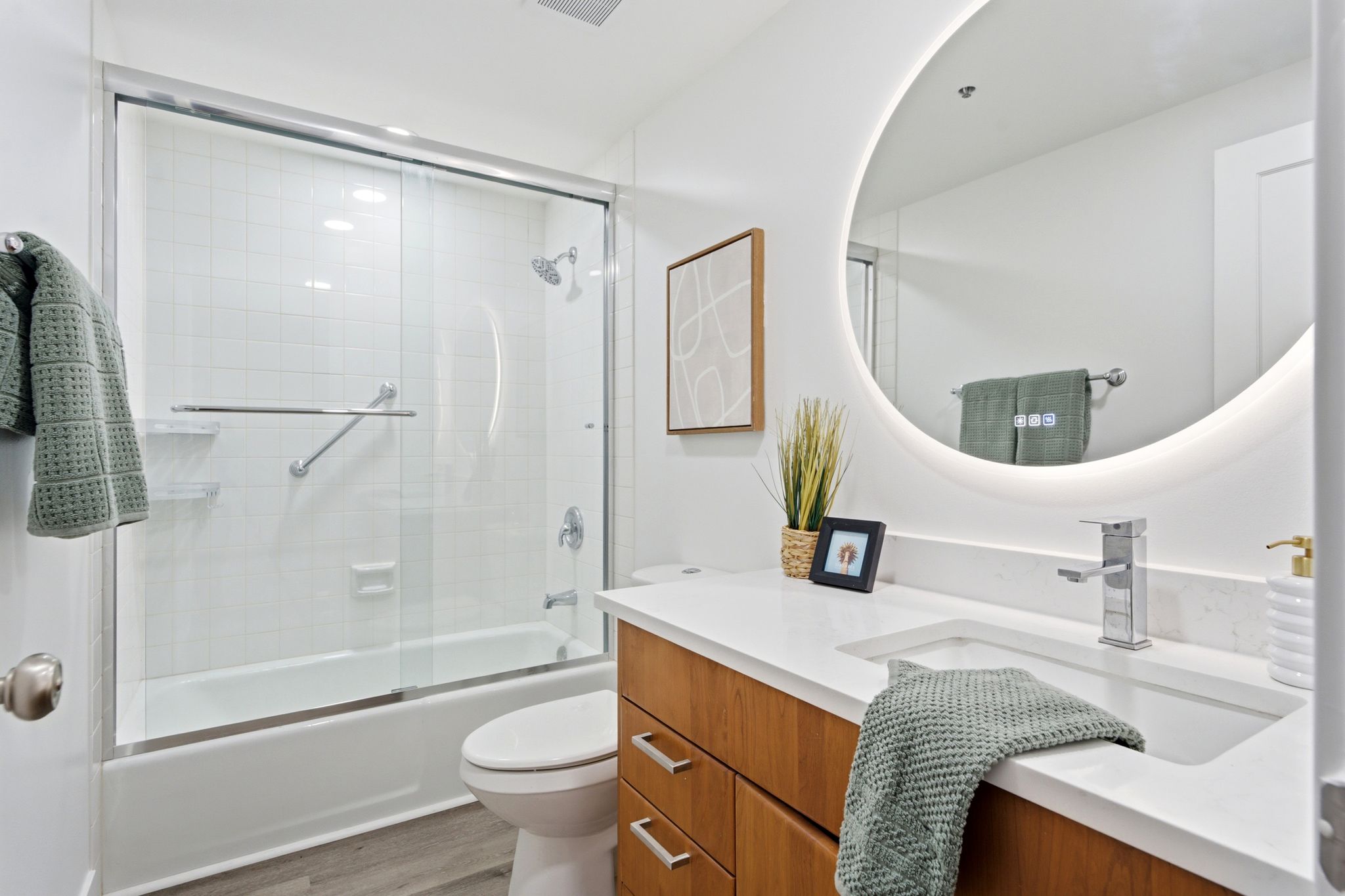 Bathroom featuring a white subway tile shower, sliding glass shower doors, a built-in bathtub, a wood-finish vanity with an integrated sink, and an illuminated circular mirror