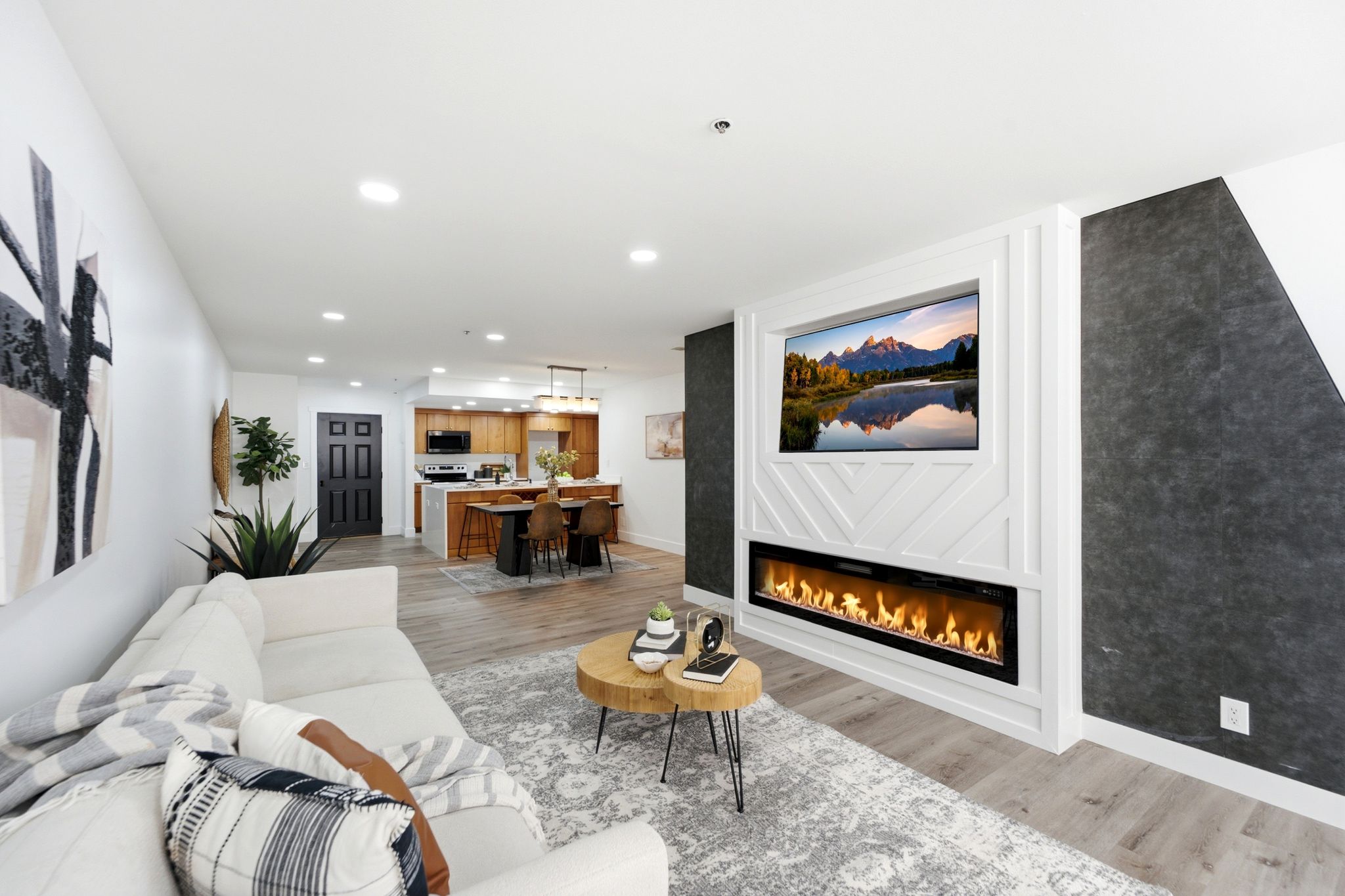 Living room featuring a built-in entertainment center with an integrated linear fireplace, wood-finish flooring, recessed lighting, and a modern kitchen visible in the background with wood cabinetry and a counter-height island