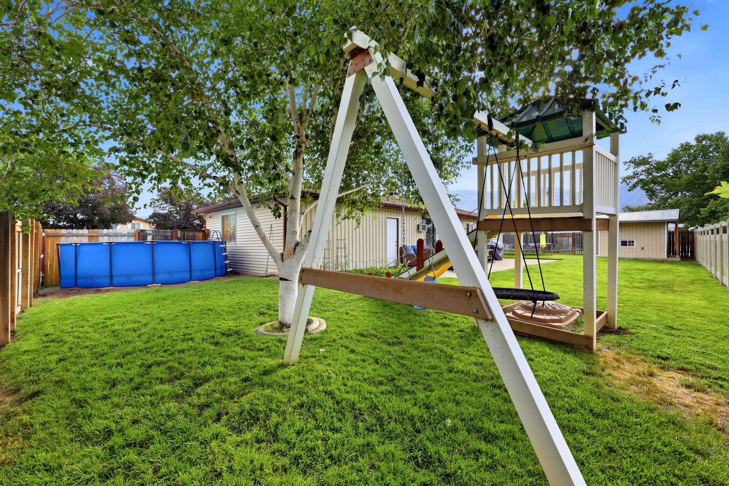 Fenced backyard featuring a play structure with swings, an above-ground pool, and mature trees