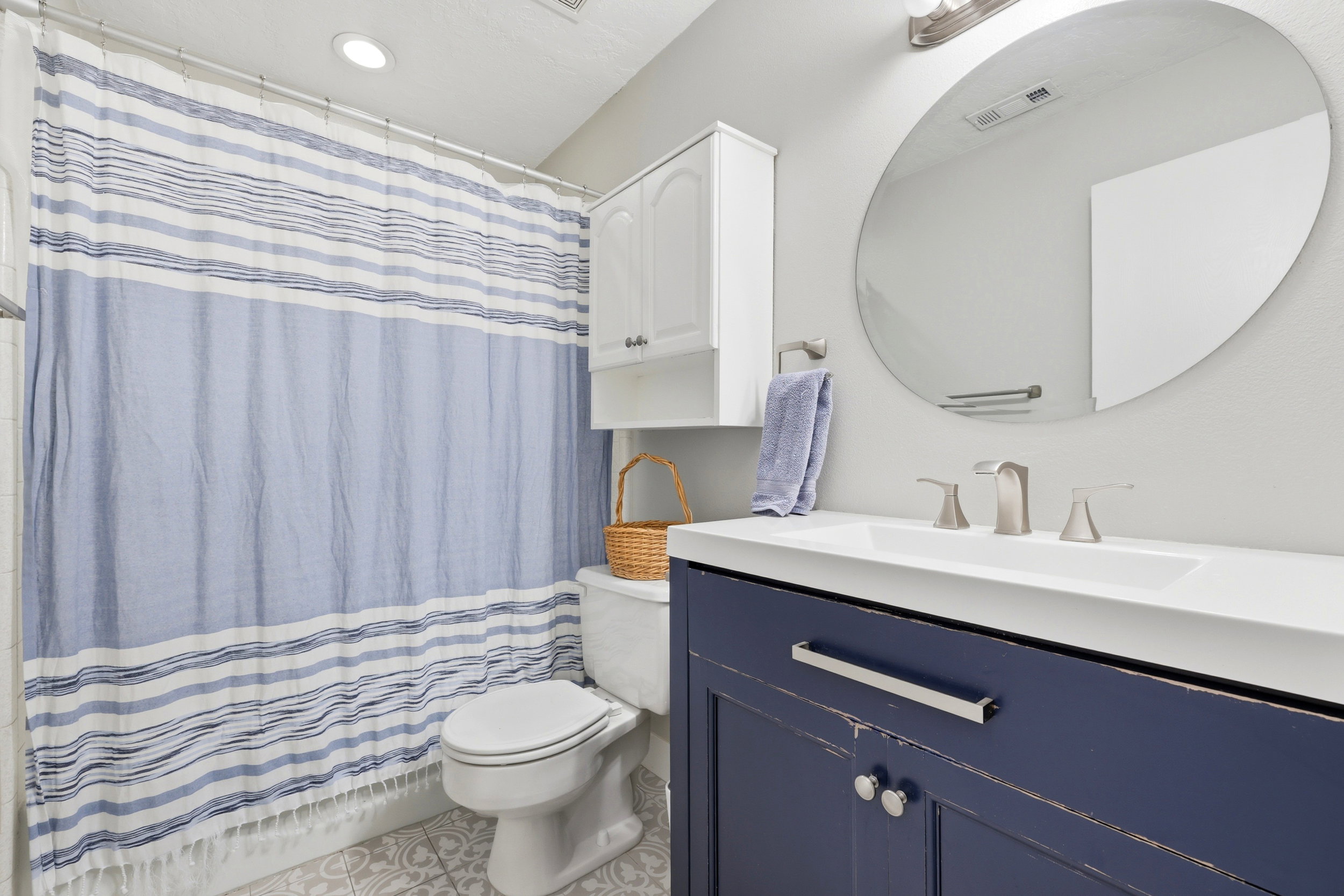 Bathroom featuring a navy vanity with brushed nickel hardware, a white integrated sink, a large oval mirror, and a white wall-mounted cabinet
