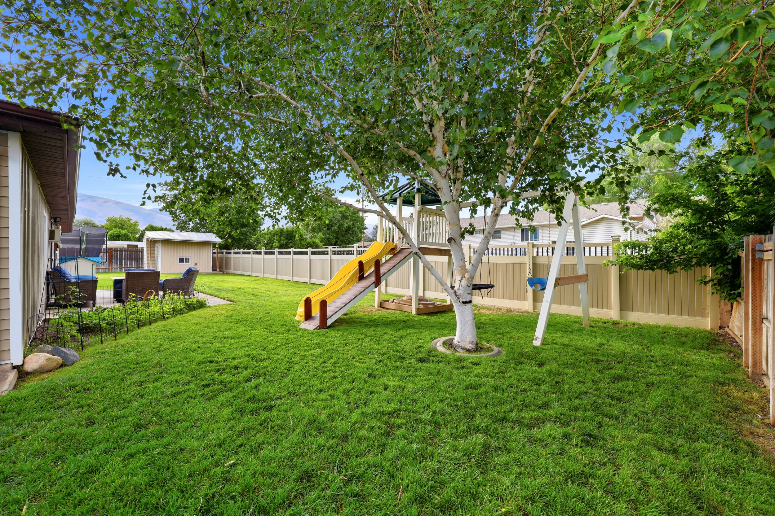 Expansive grassy backyard featuring a large mature tree, a paved patio area, a detached storage shed, and perimeter fencing