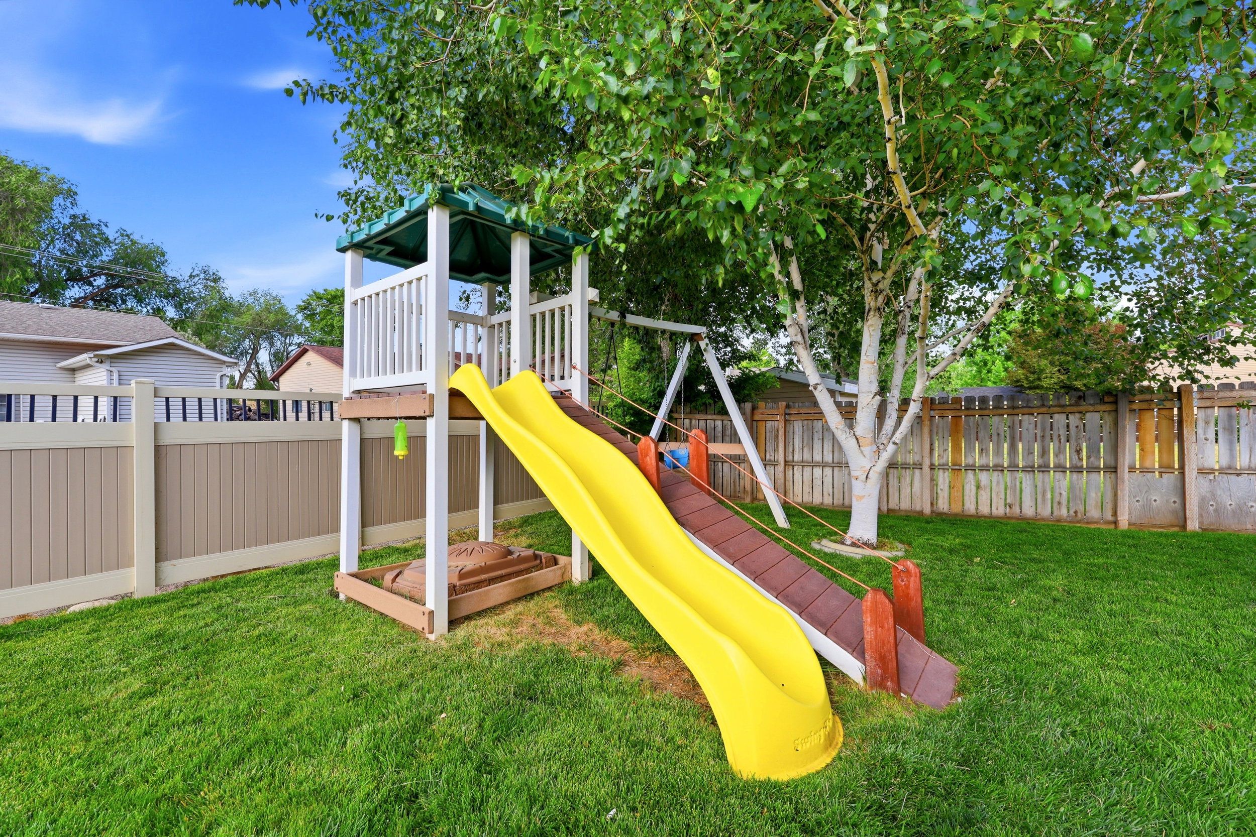 Expansive grassy backyard featuring a play structure with two slides, a swing set, and a sandbox