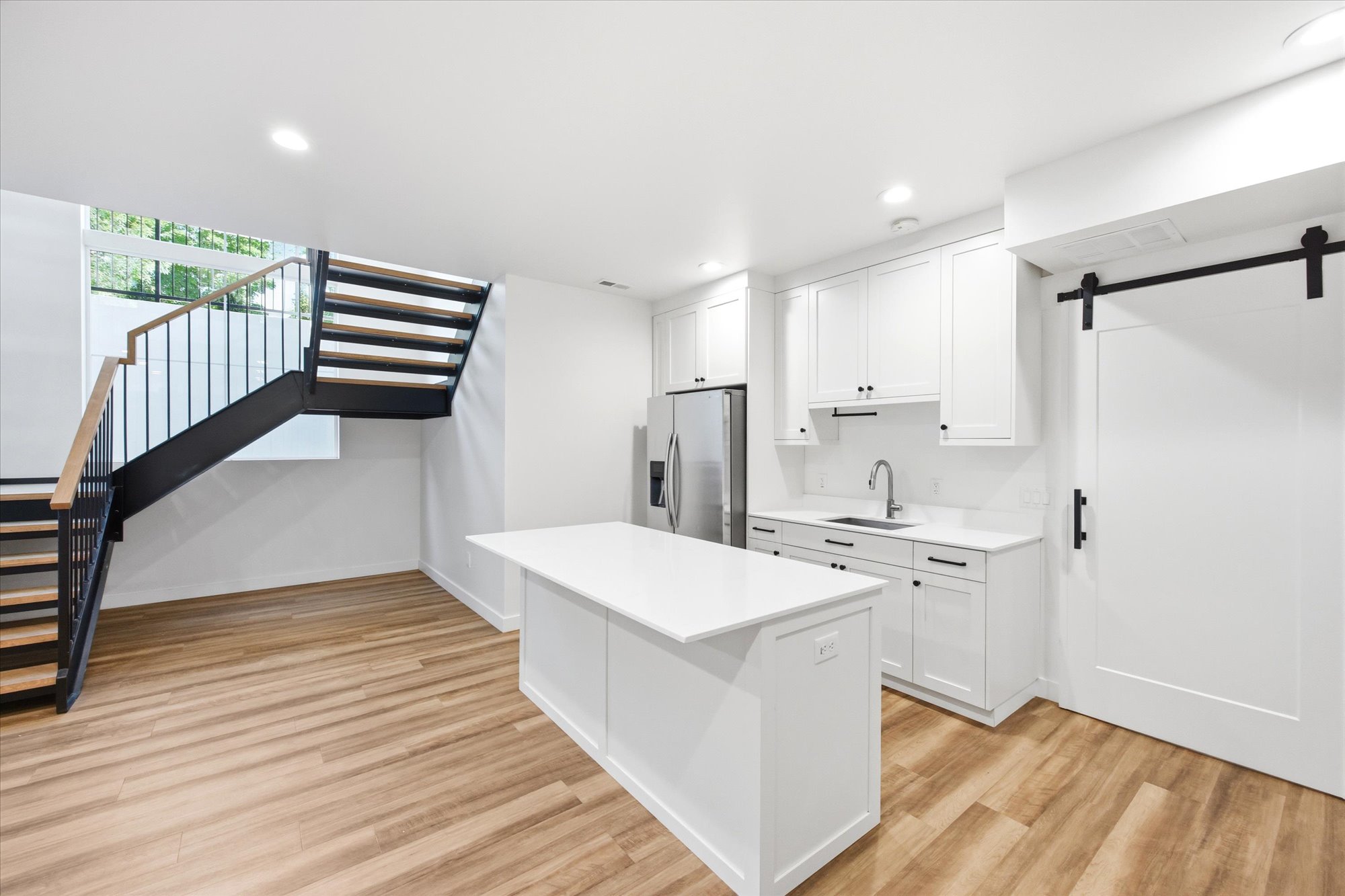 Contemporary kitchen and living area featuring a white kitchen island with quartz countertop, white shaker cabinetry, stainless steel refrigerator, and wood-finish flooring