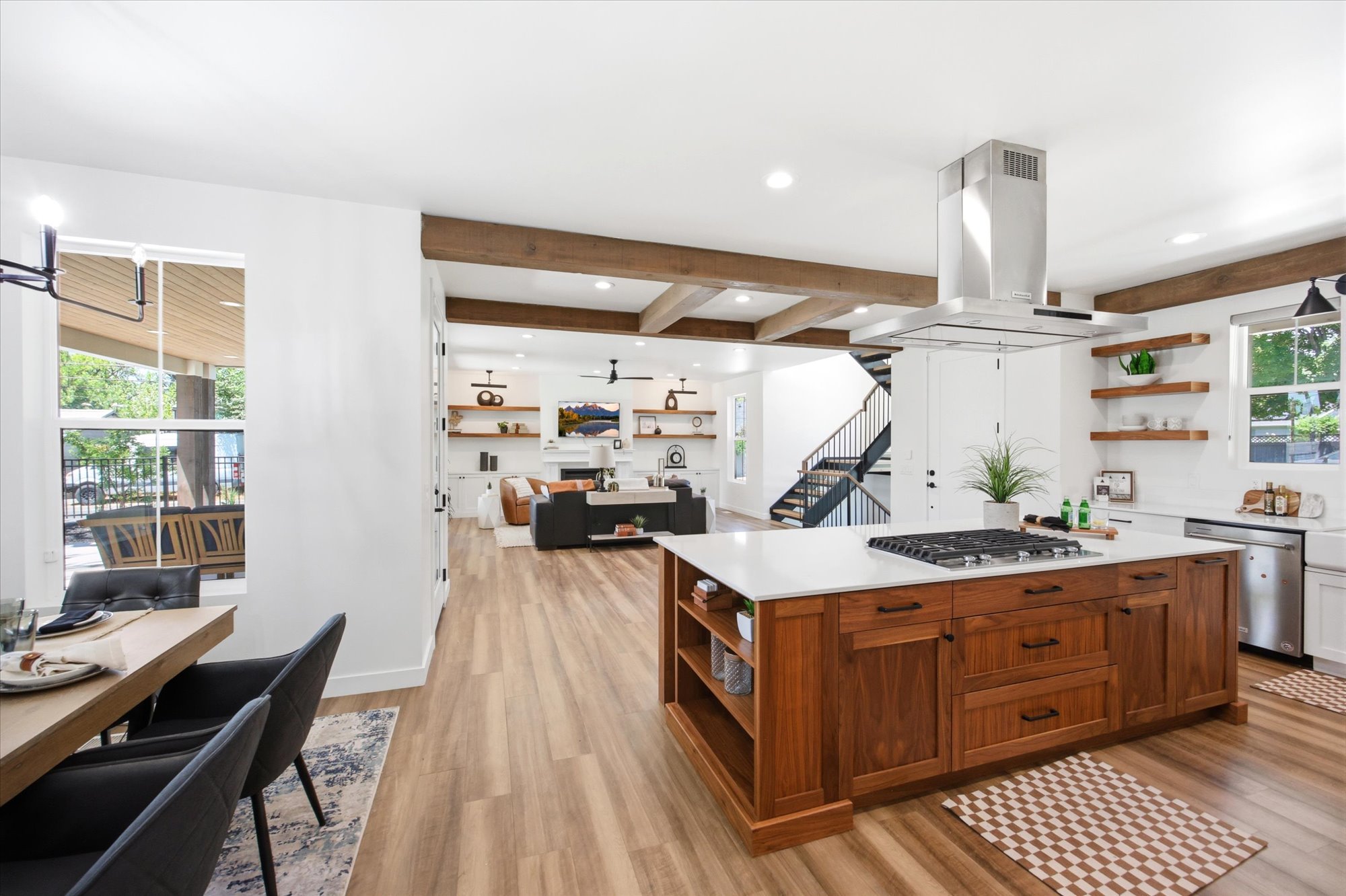 Expansive open-concept living area featuring a kitchen island with a gas cooktop, wood-finish flooring, exposed ceiling beams, a white brick fireplace, and a staircase with black metal railings