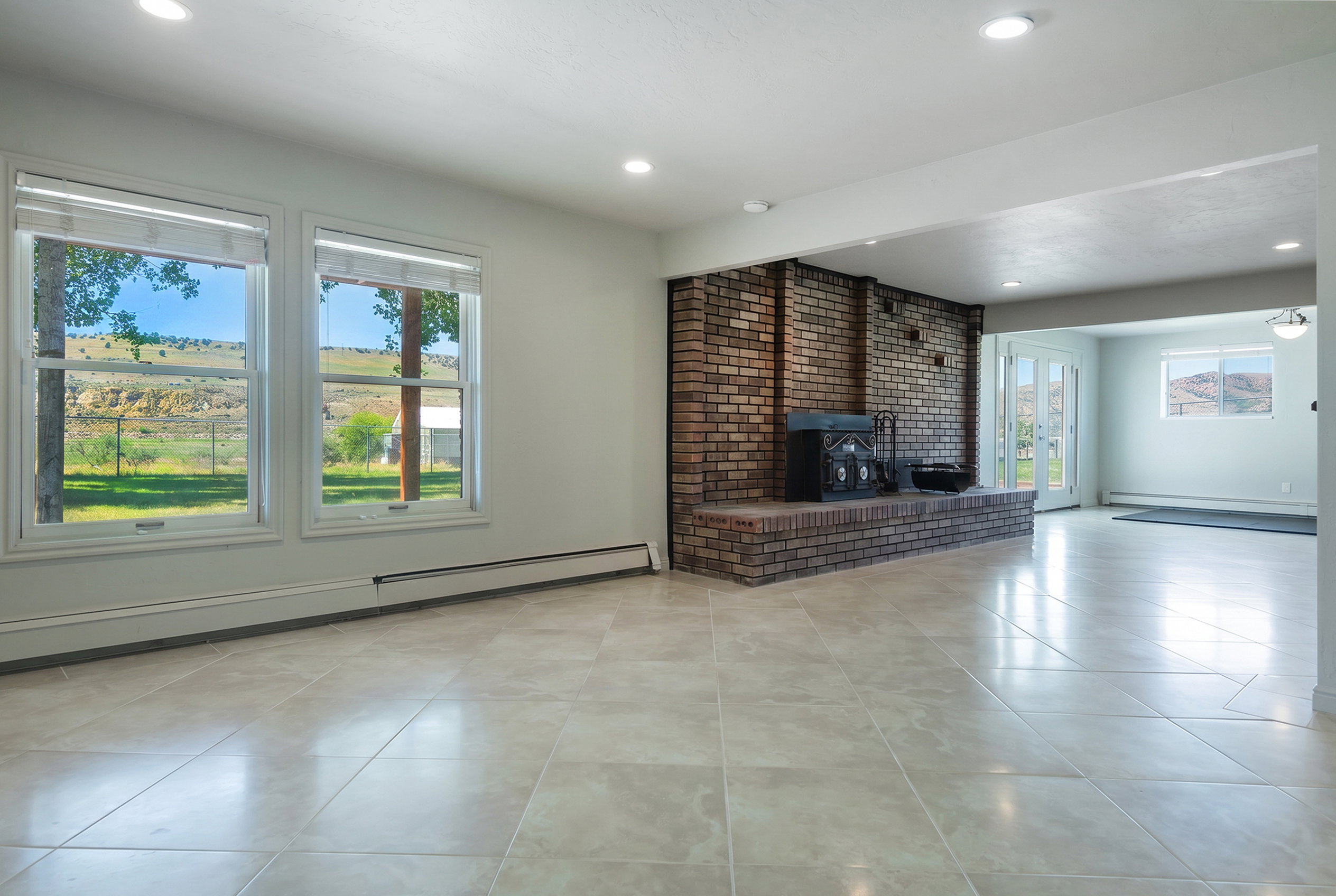 Expansive room featuring large windows, light-toned tile flooring, a brick fireplace with a hearth, recessed lighting, and French doors leading outdoors