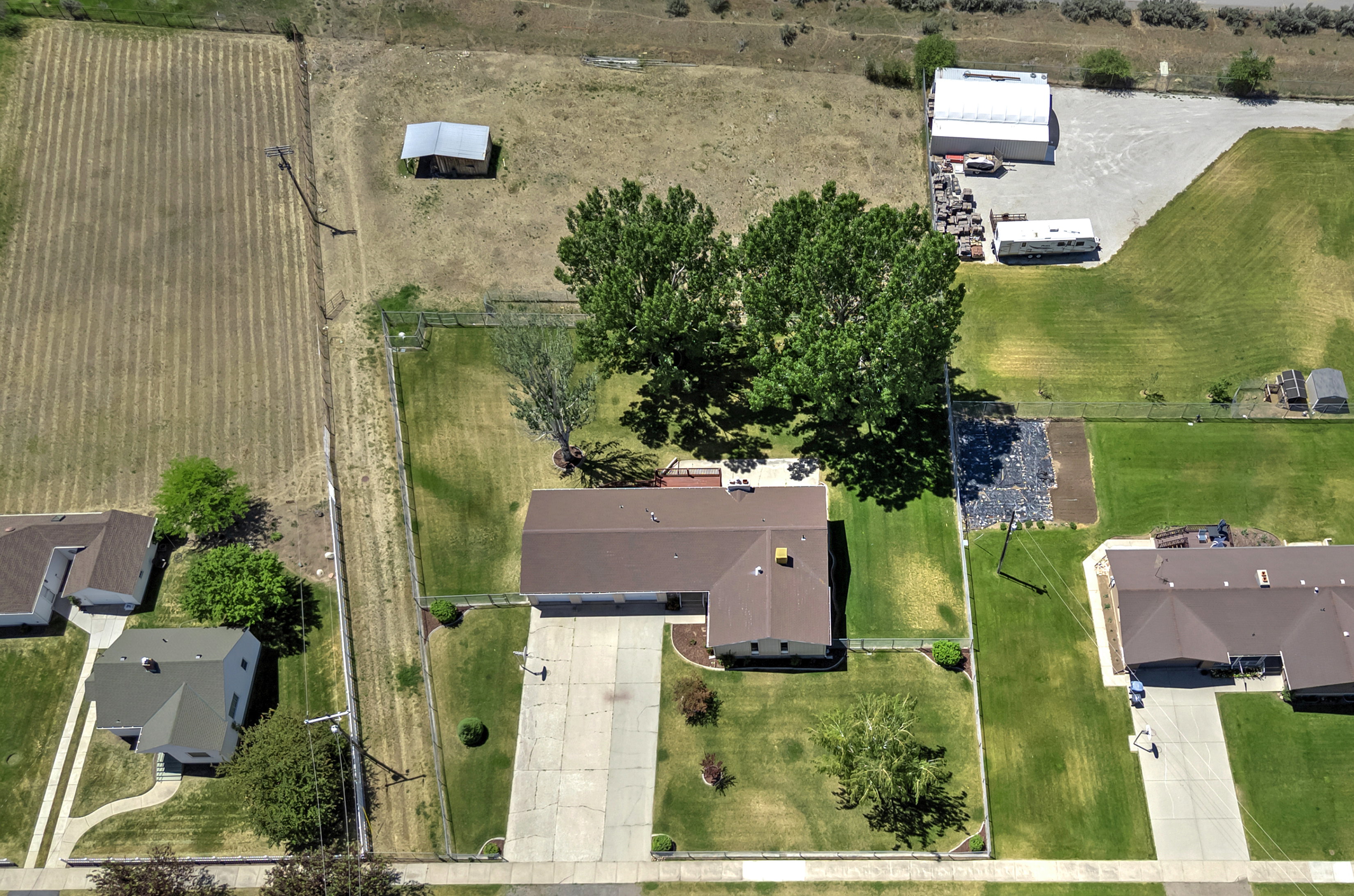 Aerial property view featuring a large green lawn, mature shade trees, a concrete driveway, and a fenced perimeter