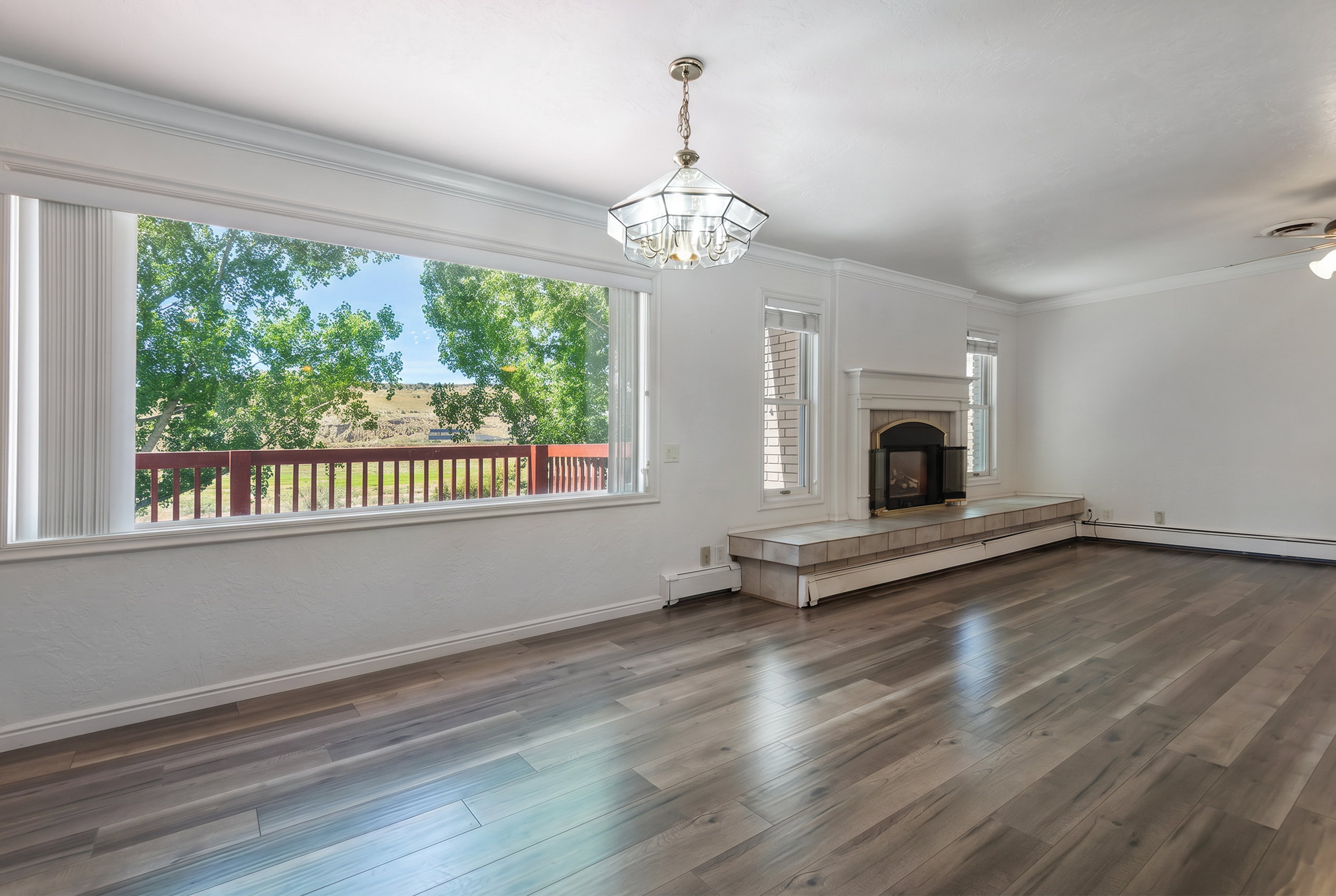 Bright living area featuring wood-finish flooring, a contemporary chandelier, a built-in fireplace with a tiled hearth, and extensive windows showcasing a balcony with a wooden railing and outdoor greenery
