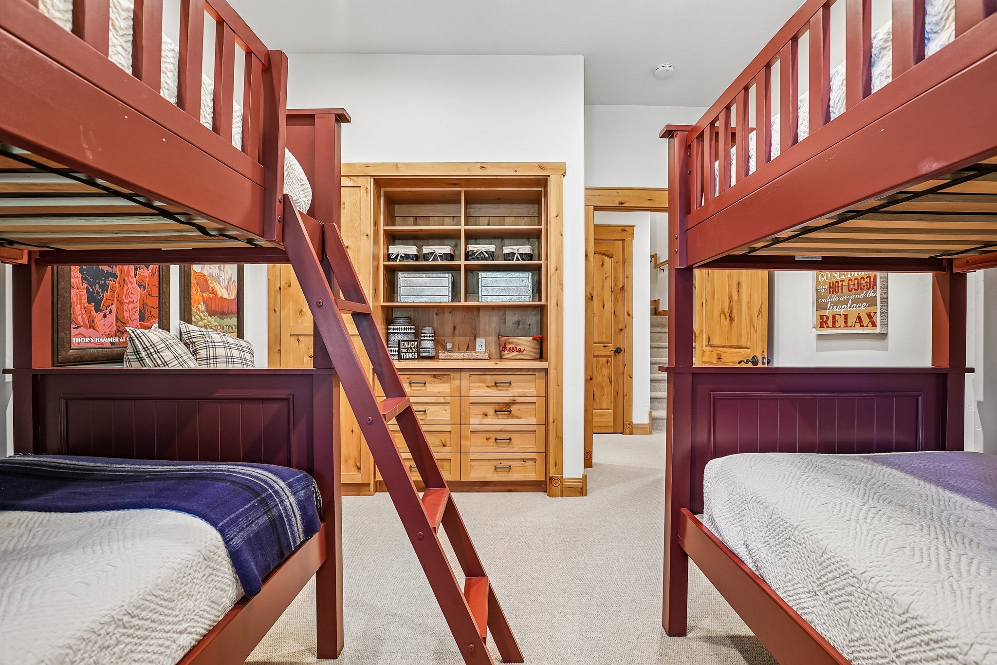 Built-in bunk beds with dark red frames, integrated ladder, natural wood-finish cabinetry with shelving, light neutral carpet, and white walls