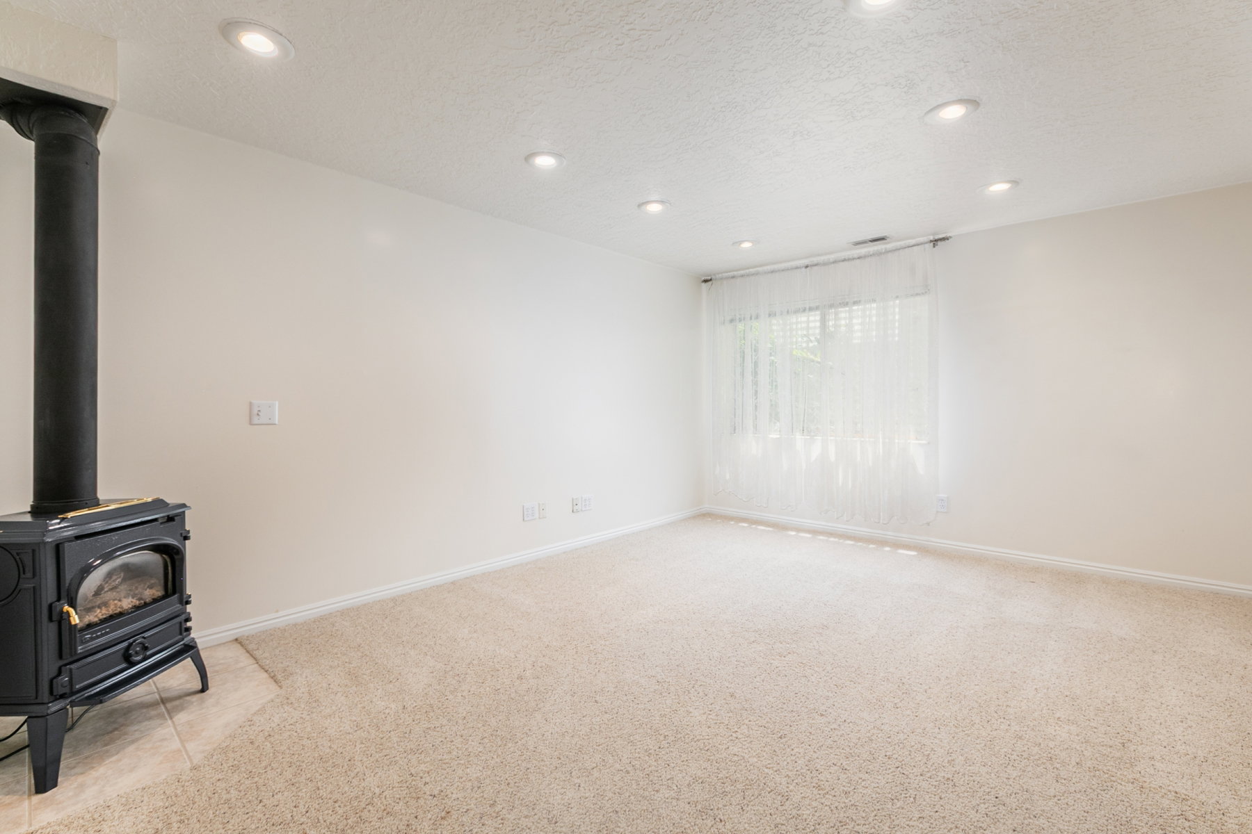 Spacious room featuring a black wood-burning stove on a tiled hearth, recessed ceiling lighting, light-toned carpeting, and a window with sheer white curtains and blinds