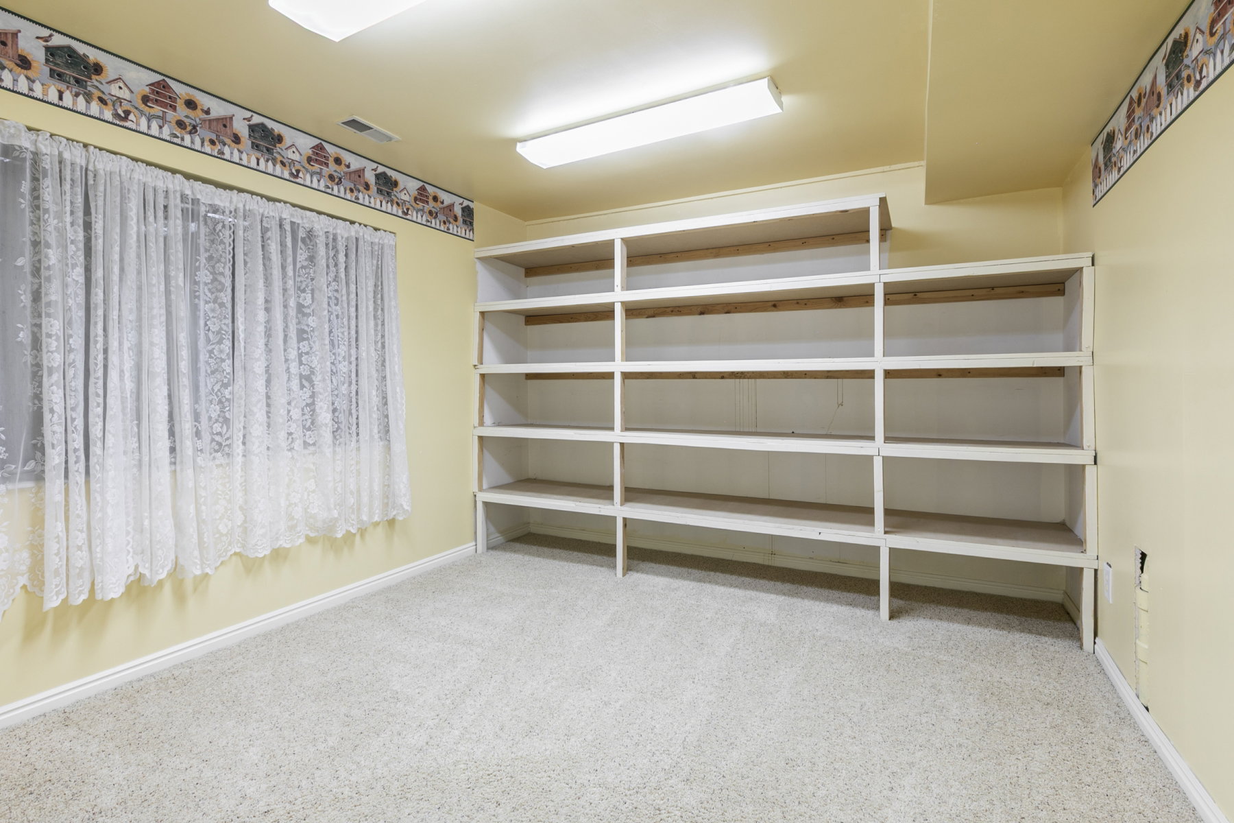 Room featuring light-colored carpeting, a window with lace curtains, and extensive built-in shelving