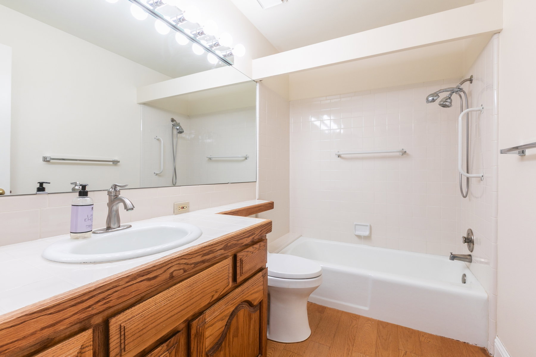 Bathroom featuring a wood-finish vanity with a white countertop and integrated sink, a large wall-mounted mirror, a tub-shower combination with white tile surround, and a dual-head shower fixture