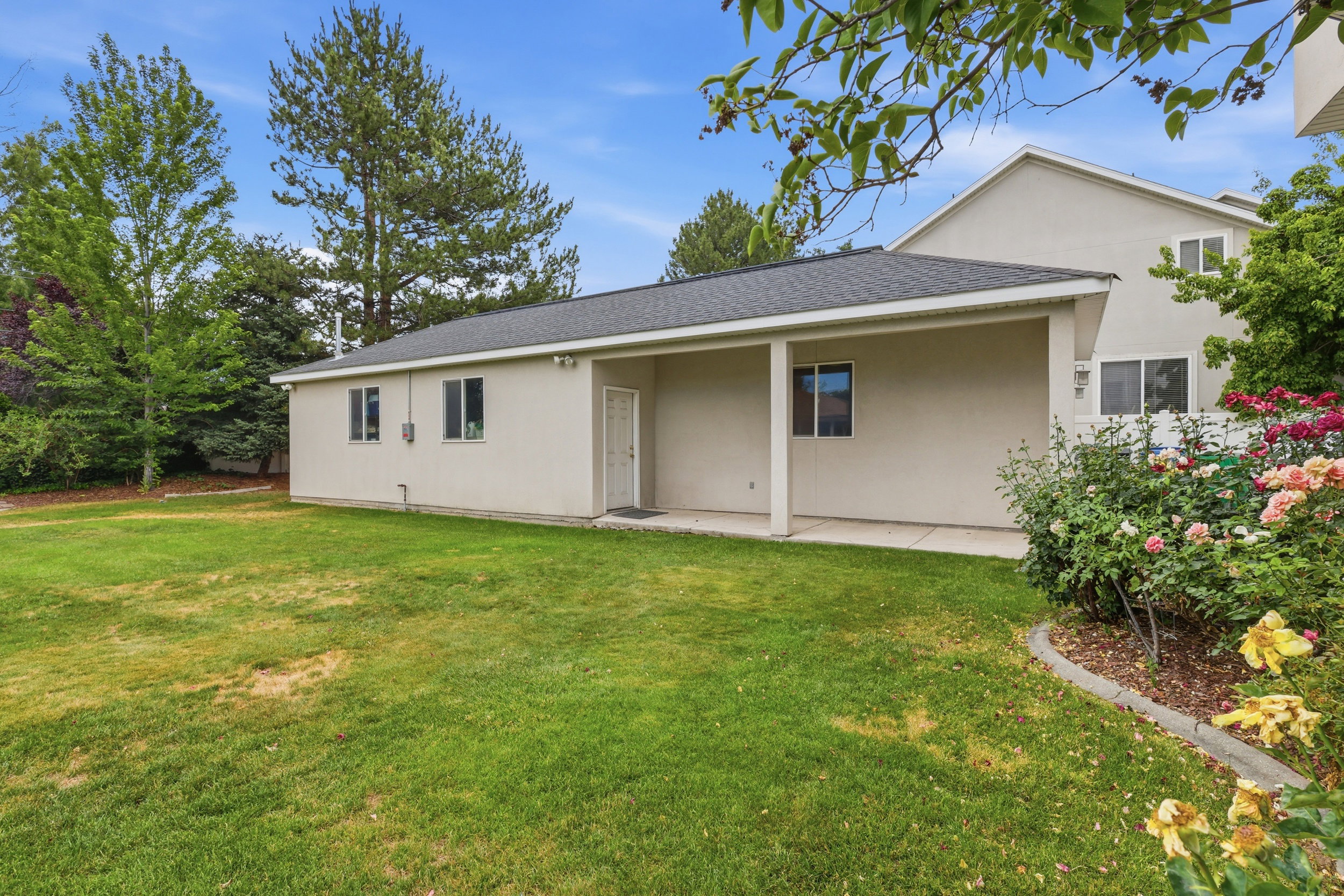 Expansive backyard featuring a covered patio, established plantings, mature trees, and a stucco exterior
