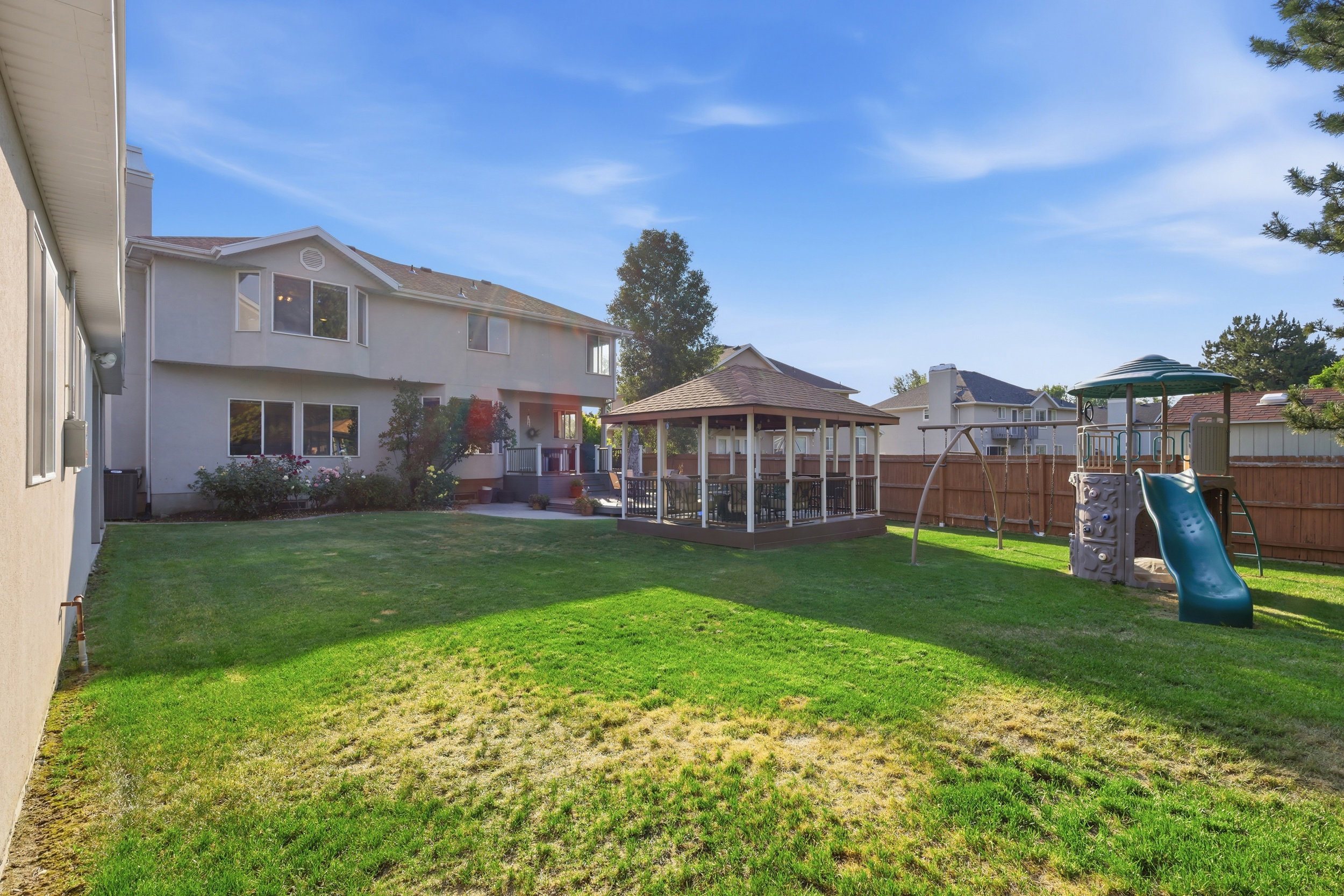 Expansive backyard featuring a lush lawn, a wood-framed gazebo with a shingled roof, a wooden privacy fence, and a multi-level play structure with a slide