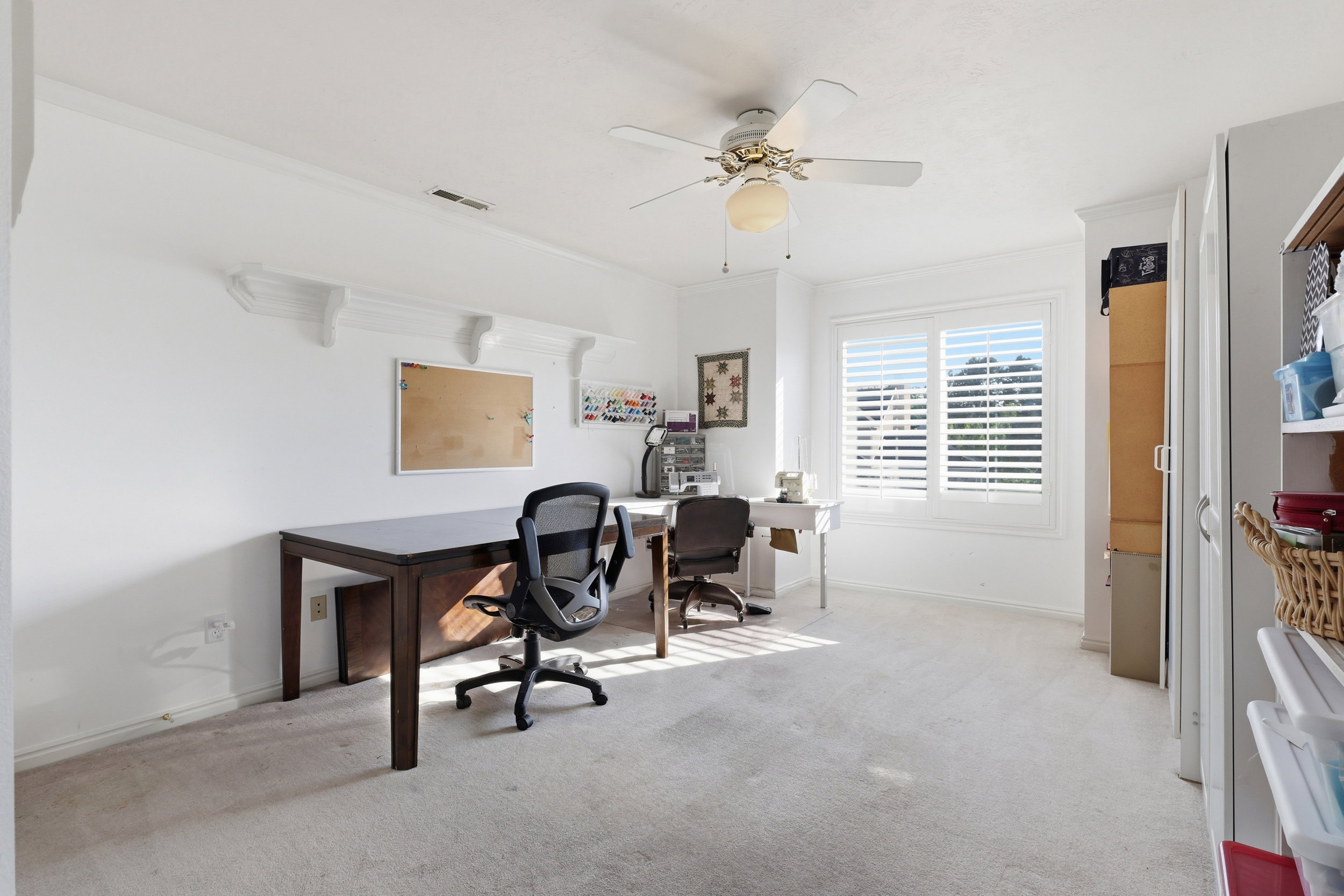 Bright room featuring a large window with white plantation shutters, a ceiling fan with light fixture, light-toned carpet flooring, and white painted walls