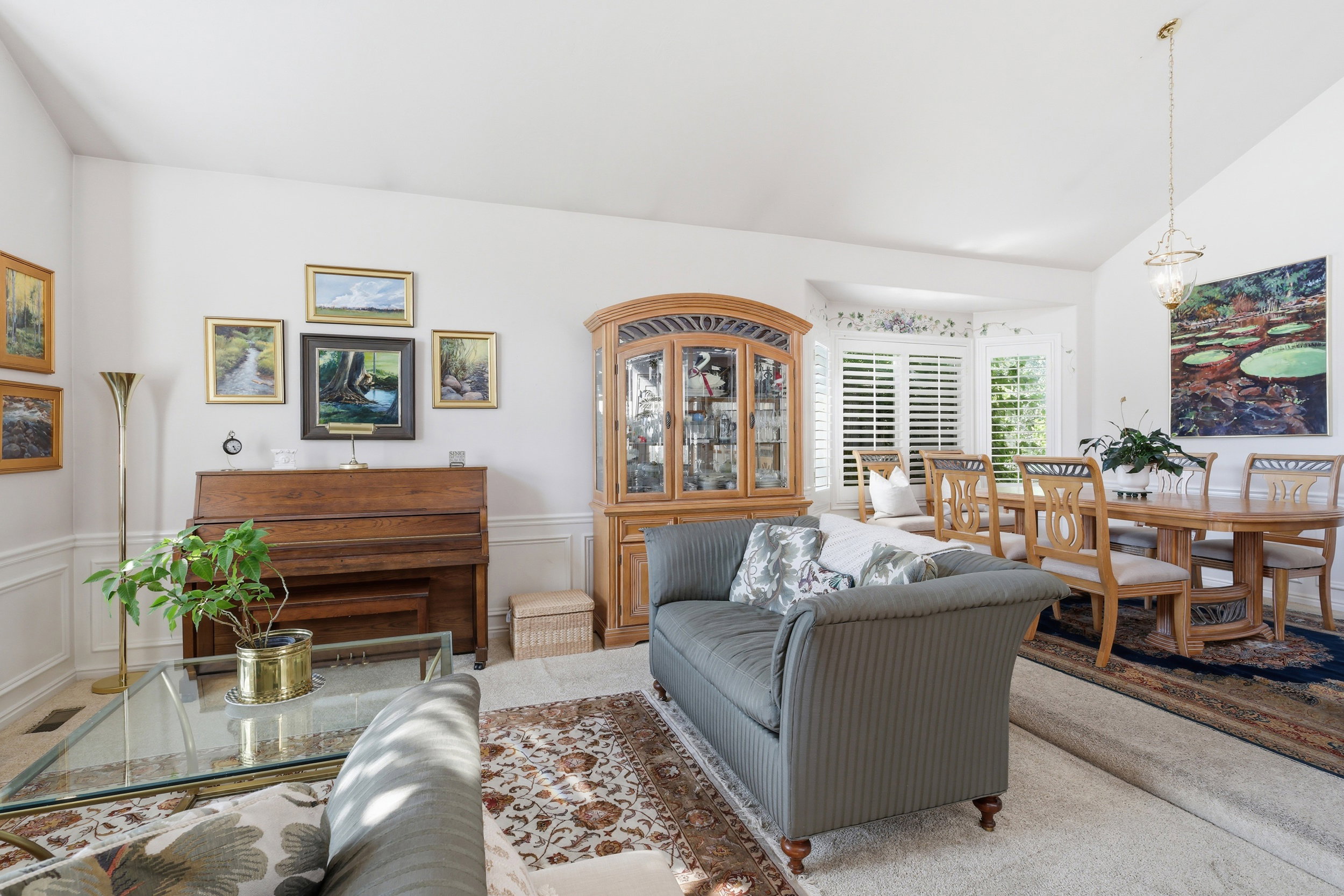 Living and dining area featuring vaulted ceilings, bay window with plantation shutters, wainscoting, and carpet flooring