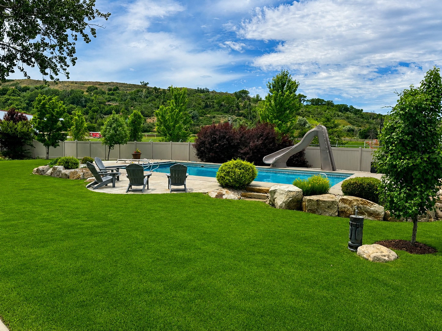 Outdoor pool area featuring a blue-tiled swimming pool with an attached slide, a surrounding stone patio, and a privacy fence
