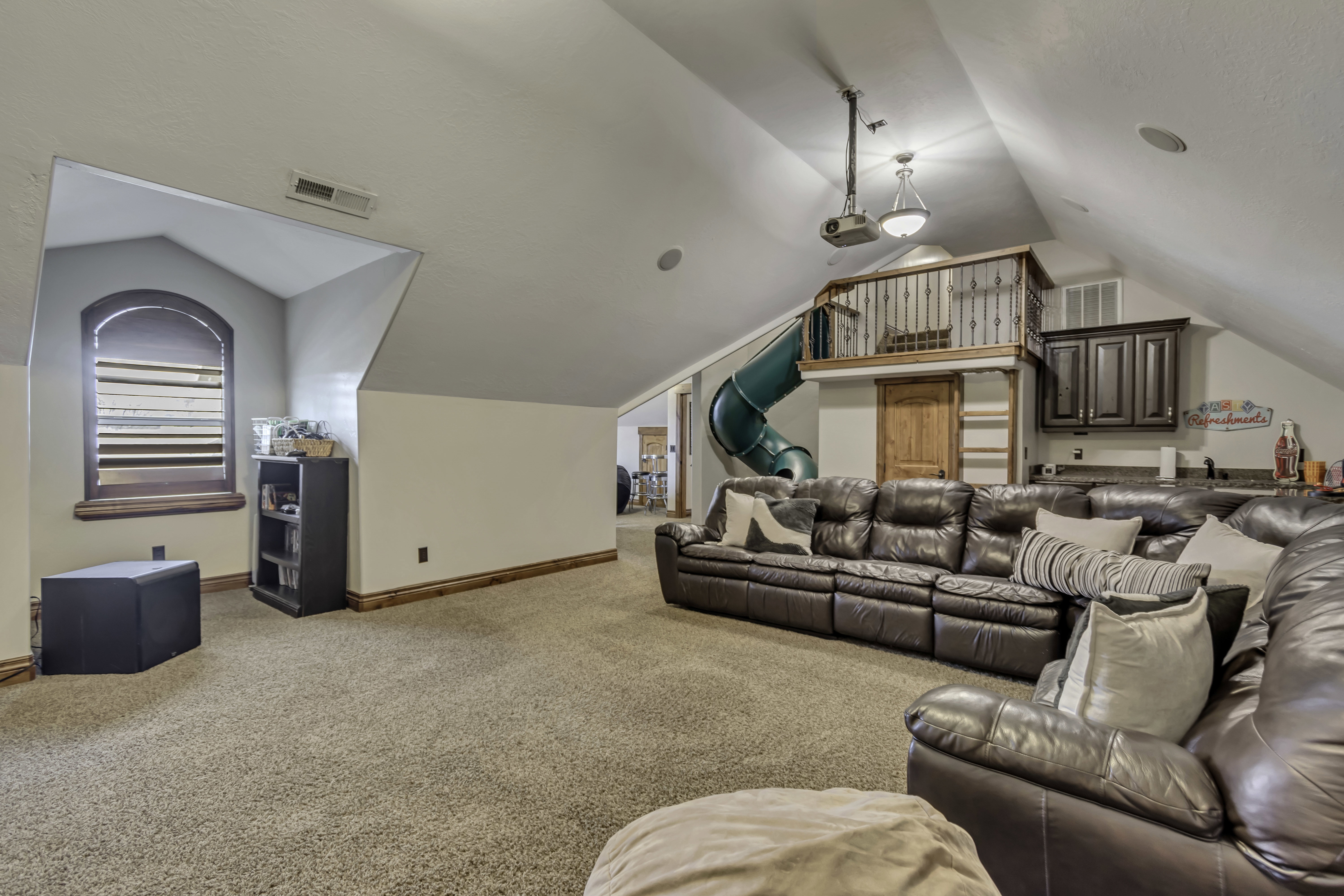 Carpeted entertainment area featuring a built-in green slide, a raised loft with metal railing, an arched window, and recessed lighting