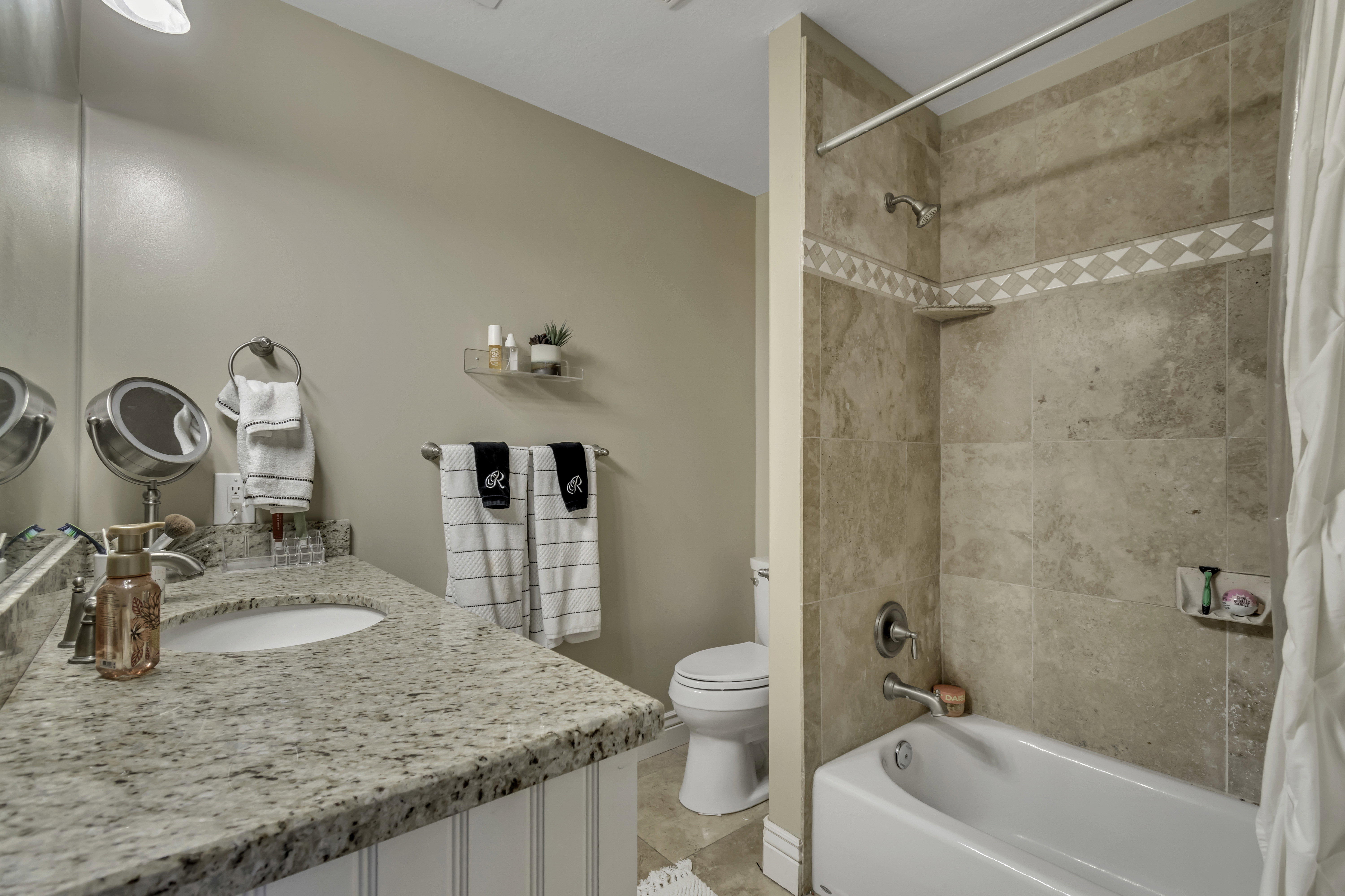 Bathroom featuring a granite countertop vanity with beadboard paneling, a full mirror, and a built-in tub with stone-look tile surround and decorative mosaic accent