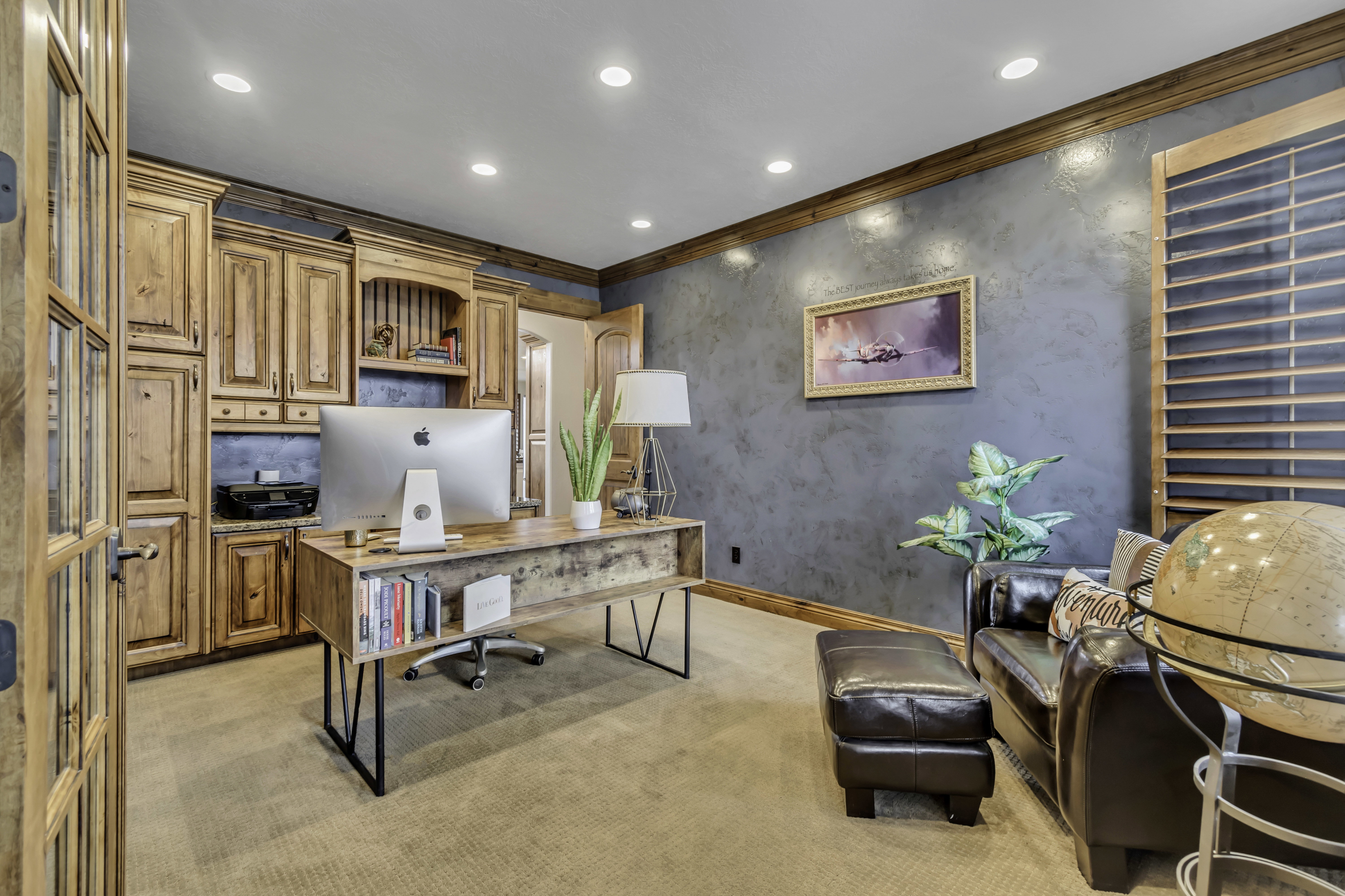Dedicated office space featuring extensive built-in wooden cabinetry, a textured accent wall, recessed lighting, and a large window with wooden shutters
