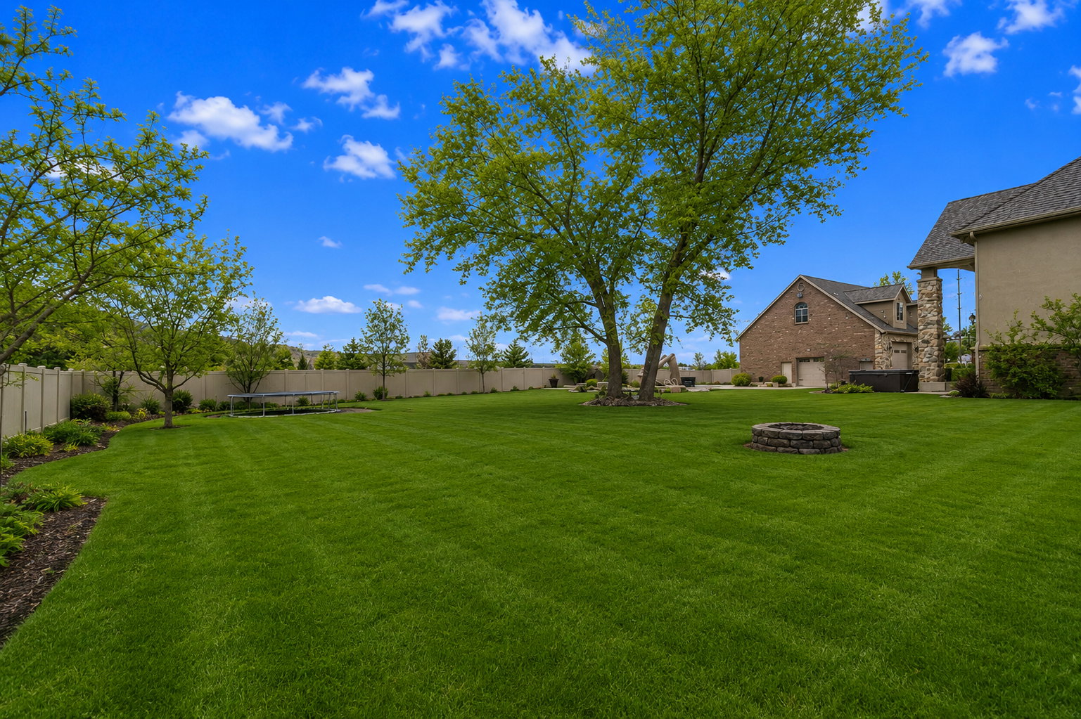 Expansive green lawn with mature trees, a stone fire pit, and a privacy fence