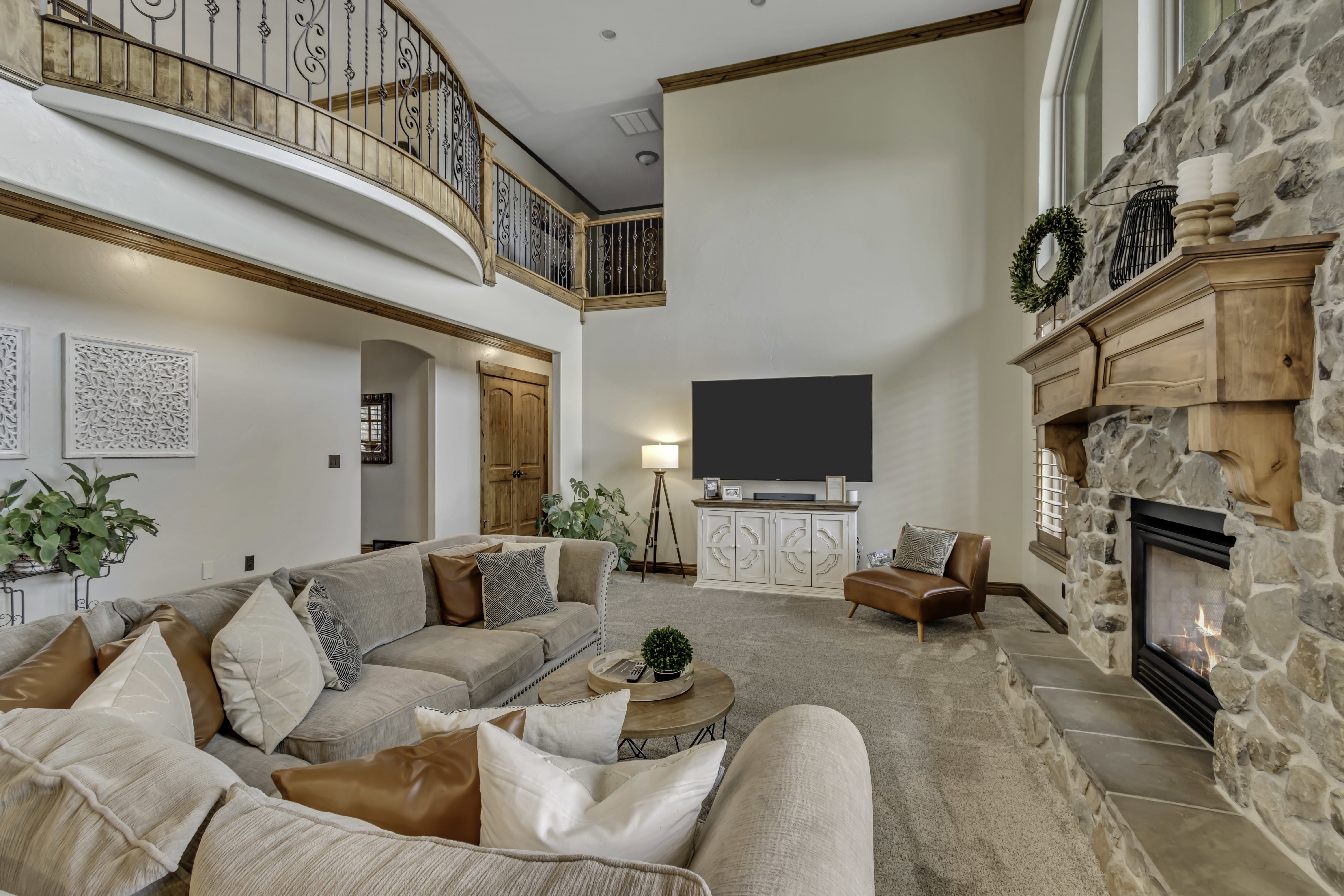 Spacious living area featuring a floor-to-ceiling stone fireplace with a wood mantle, high ceilings, and a curved second-story balcony with decorative iron railings