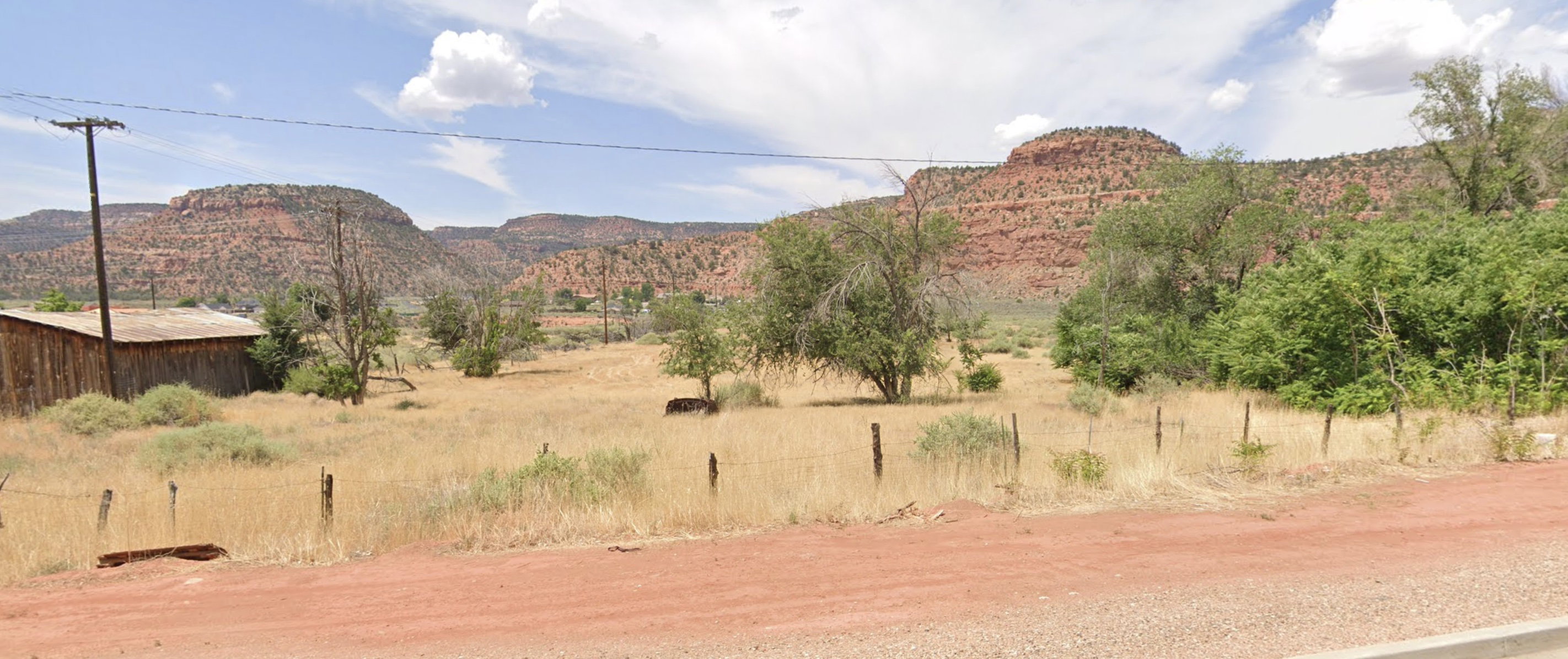 Expansive natural landscape with arid vegetation and mature trees, featuring distant mesa formations
