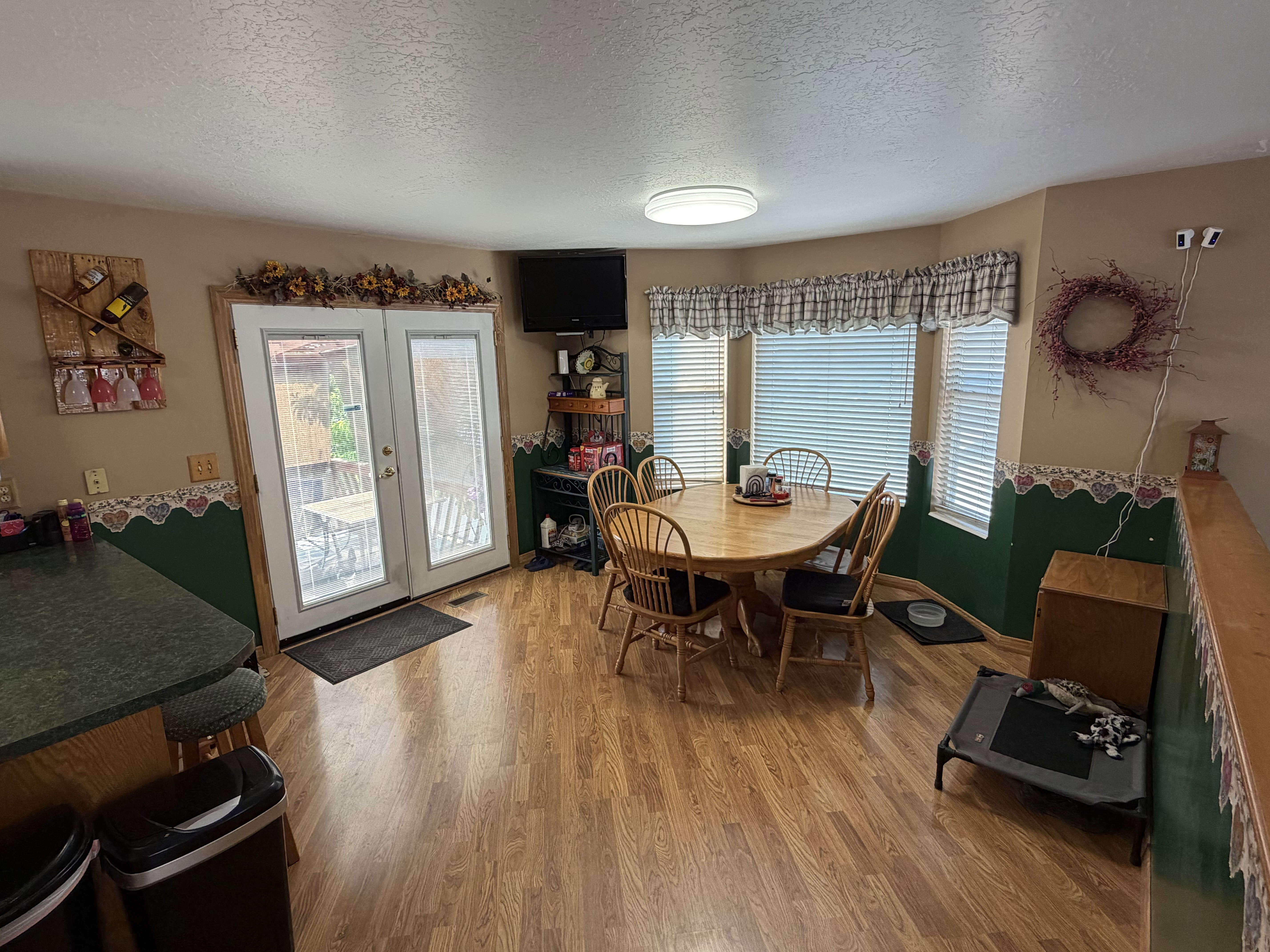 Open-concept dining area featuring wood-finish flooring, a bay window with blinds, and double glass-paneled doors leading to an outdoor space