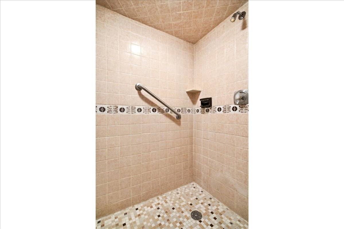 Tiled shower stall featuring mosaic floor tiling, neutral wall tiling, an accent tile band, and a chrome grab bar