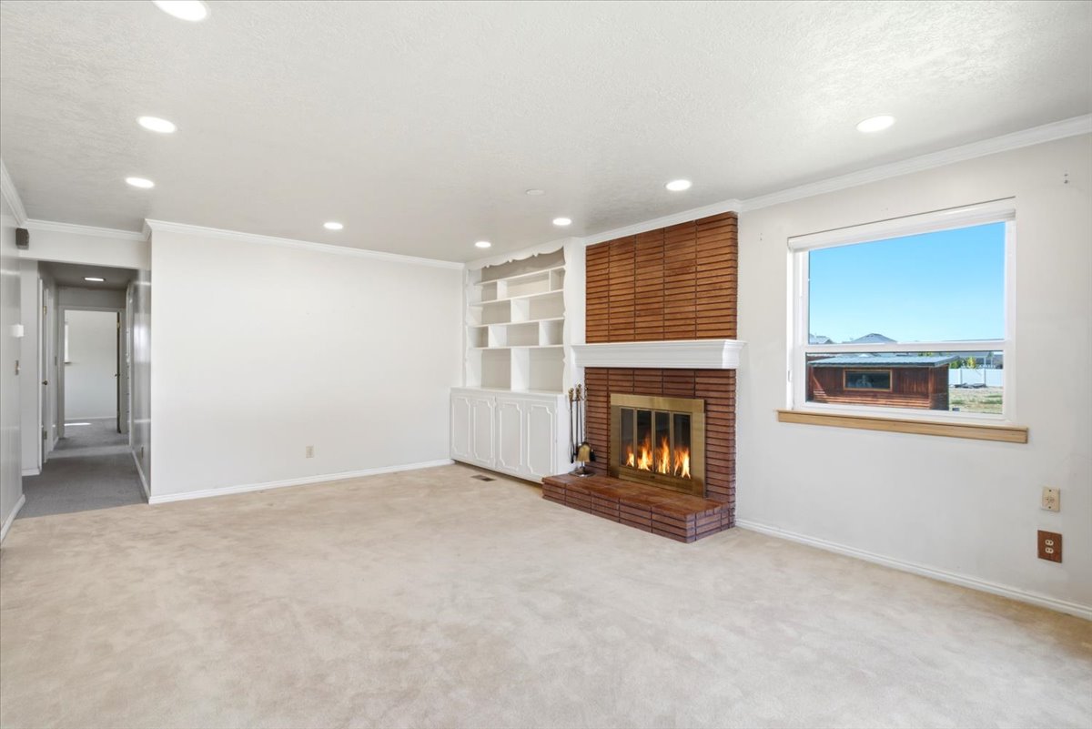 Living space featuring a brick fireplace with a white mantel, built-in shelving, recessed ceiling lights, a window, and light-toned carpet flooring