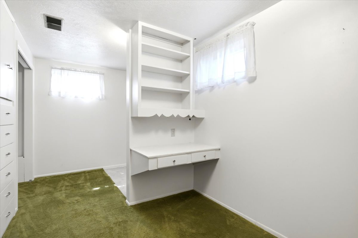 Built-in desk with shelving and integrated drawers, a window with sheer curtains, and green carpet flooring