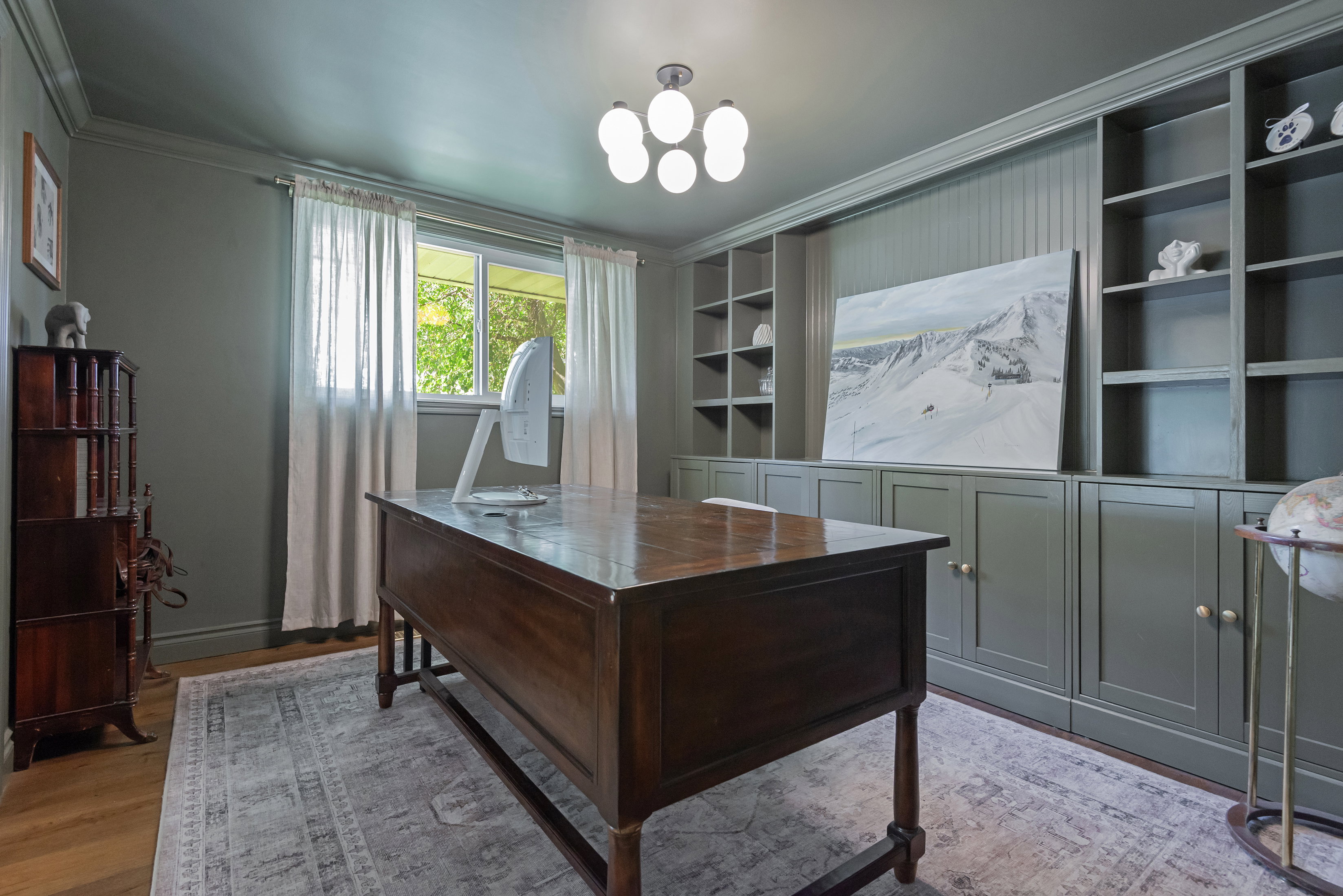 Dedicated office space featuring extensive built-in shelving and cabinetry, recessed paneling, wood-finish flooring, and a multi-bulb ceiling fixture