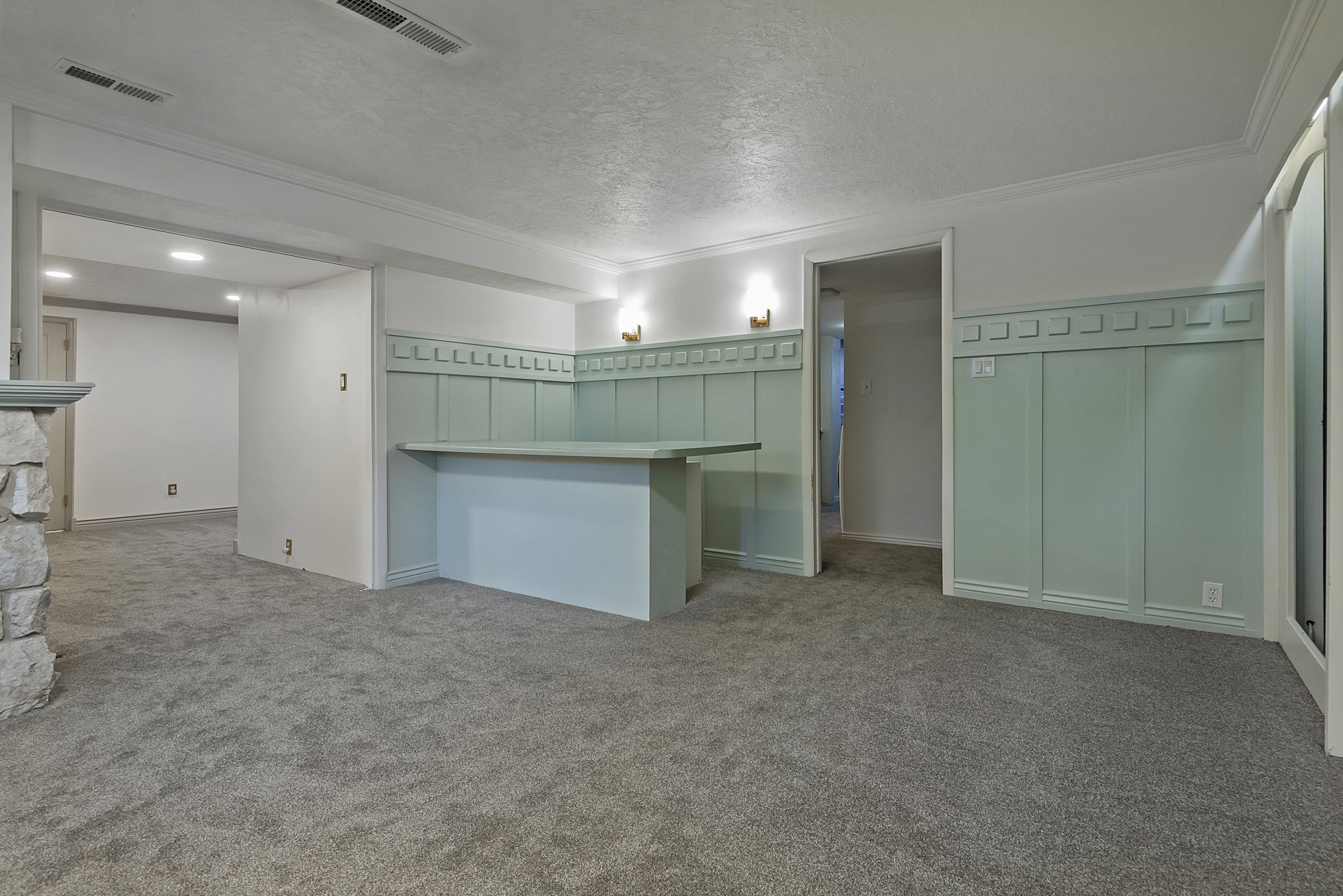 Spacious carpeted room featuring a built-in bar with light green paneling, decorative wall sconces, and crown molding