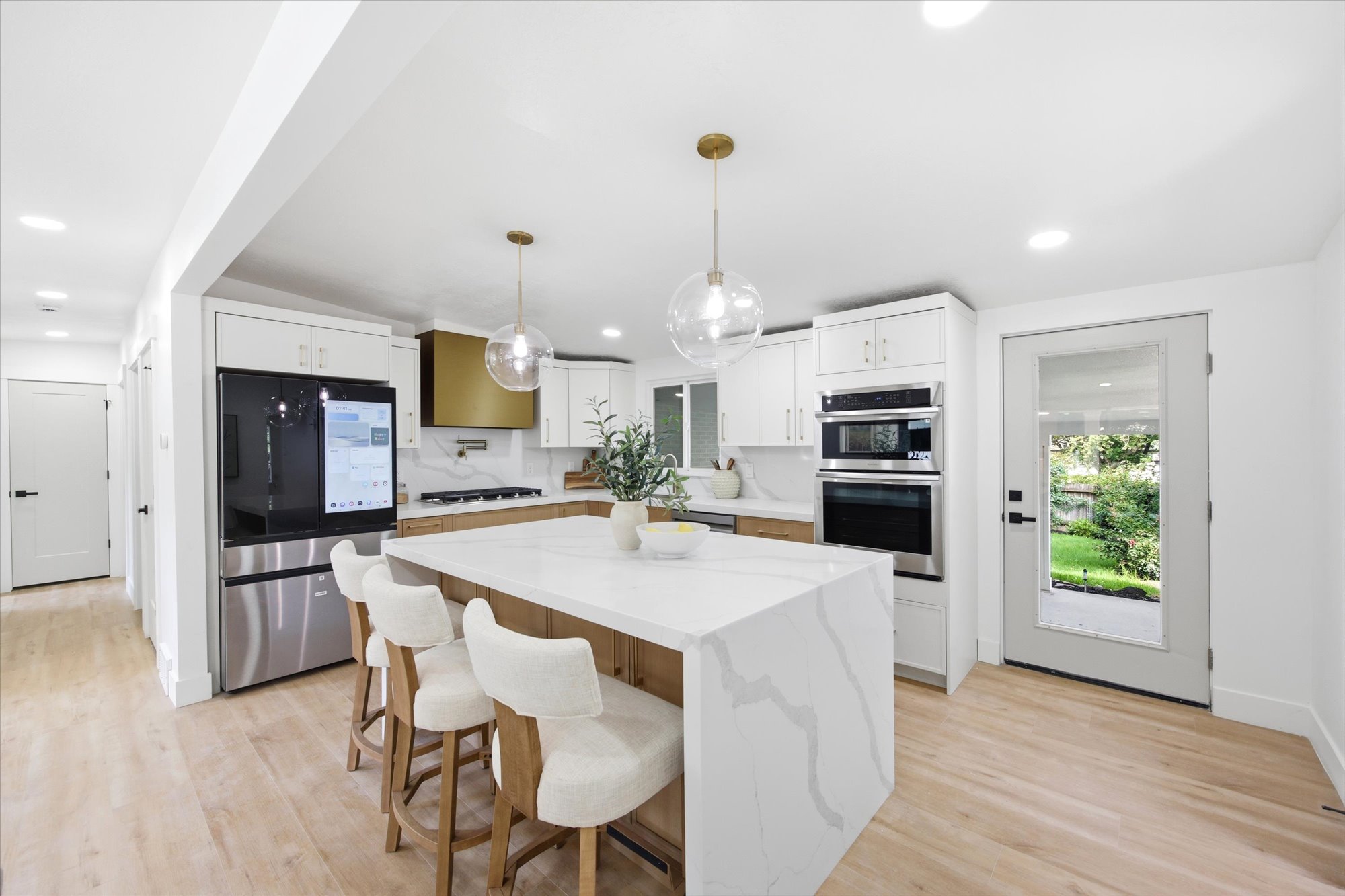 Kitchen featuring a waterfall island with stone countertop, stainless steel appliances, white cabinetry, wood-finish flooring, and a gold-tone range hood