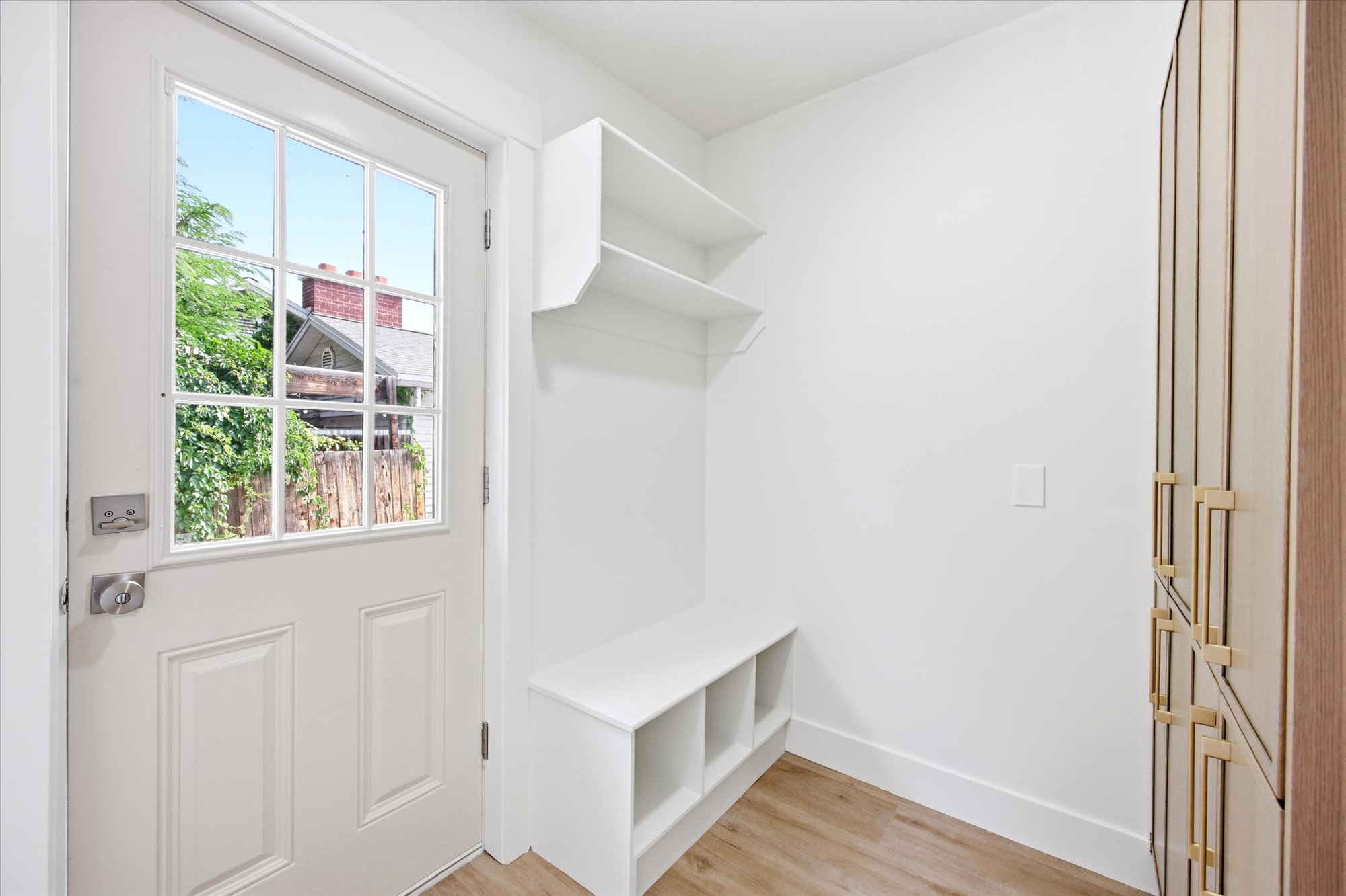Bright white entryway featuring a glass-paned door, built-in bench with cubbies, upper shelving, and wood-finish flooring