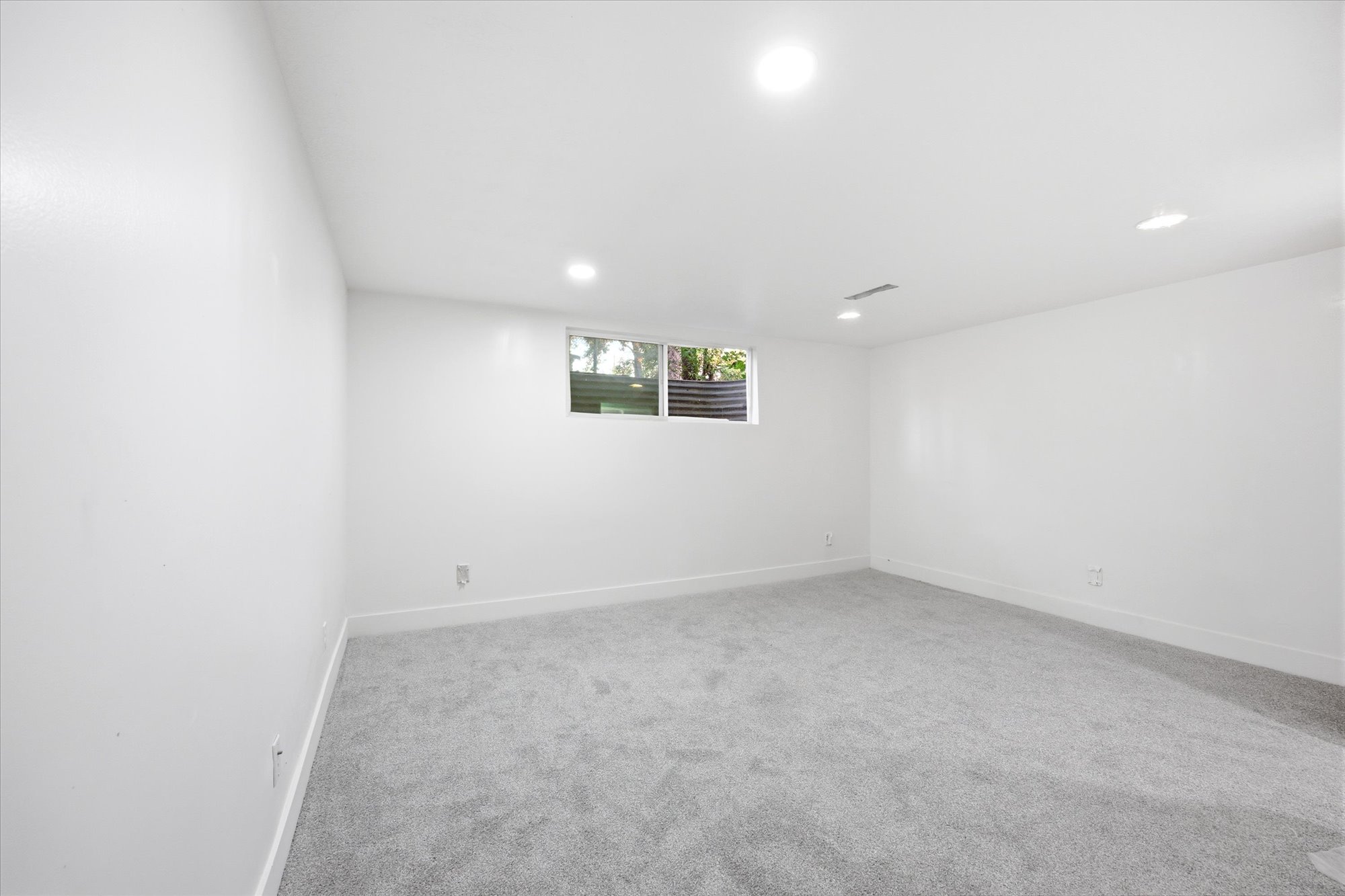 Spacious interior room featuring light gray carpet flooring, white walls, recessed ceiling lighting, and a single window