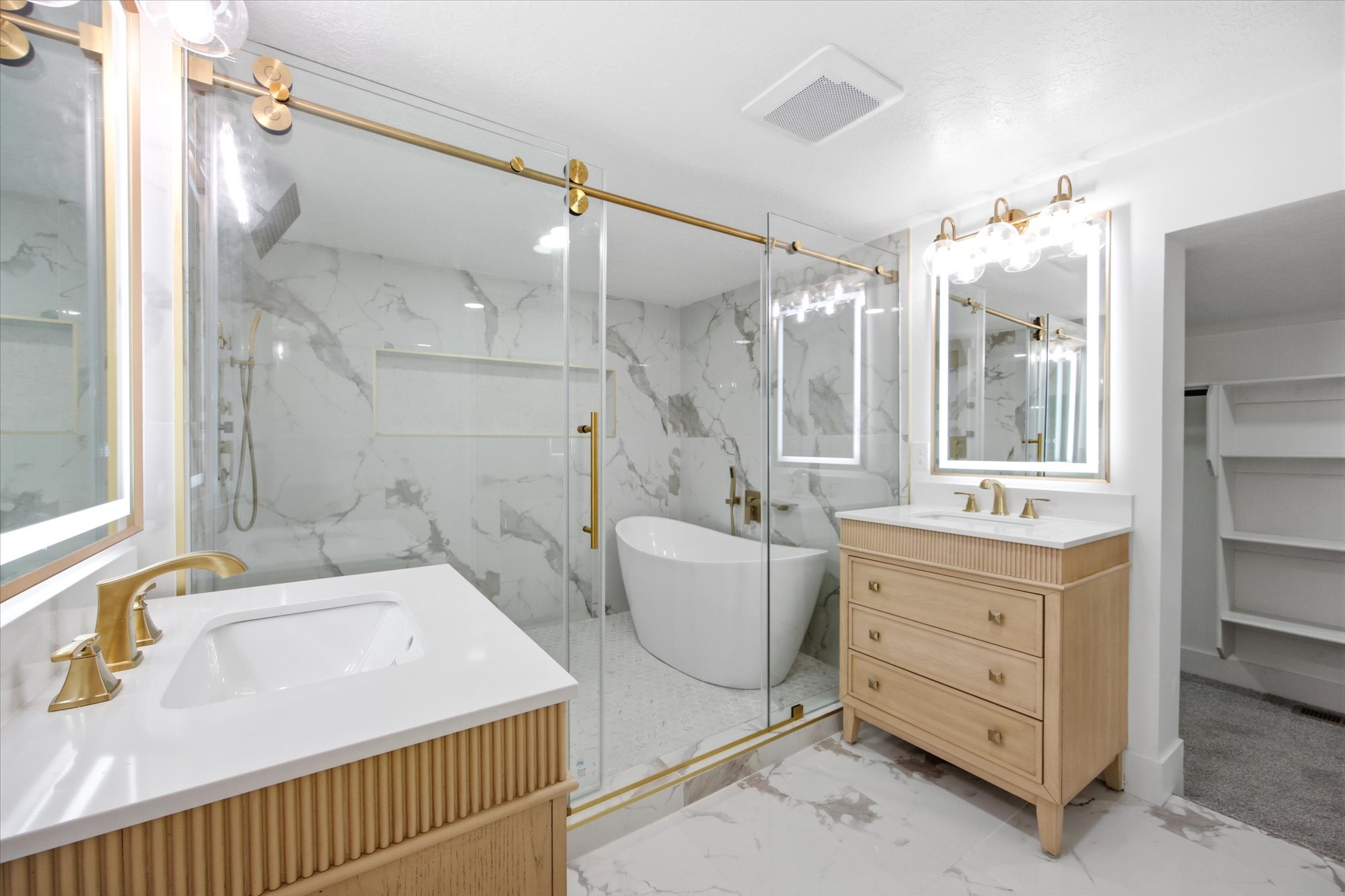 Contemporary bathroom featuring a freestanding tub, glass-enclosed shower, dual vanity sinks, illuminated mirrors, and gold-finish fixtures