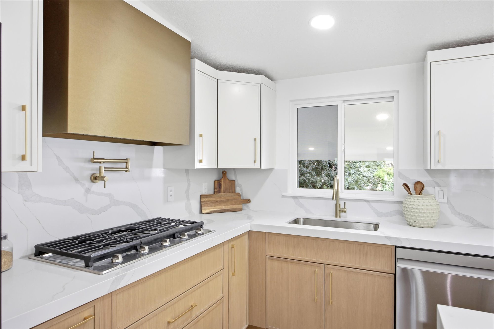 Modern kitchen featuring a brass range hood, stainless steel gas cooktop, white upper cabinetry, natural wood-finish lower cabinetry, and white countertops with a marble-effect backsplash
