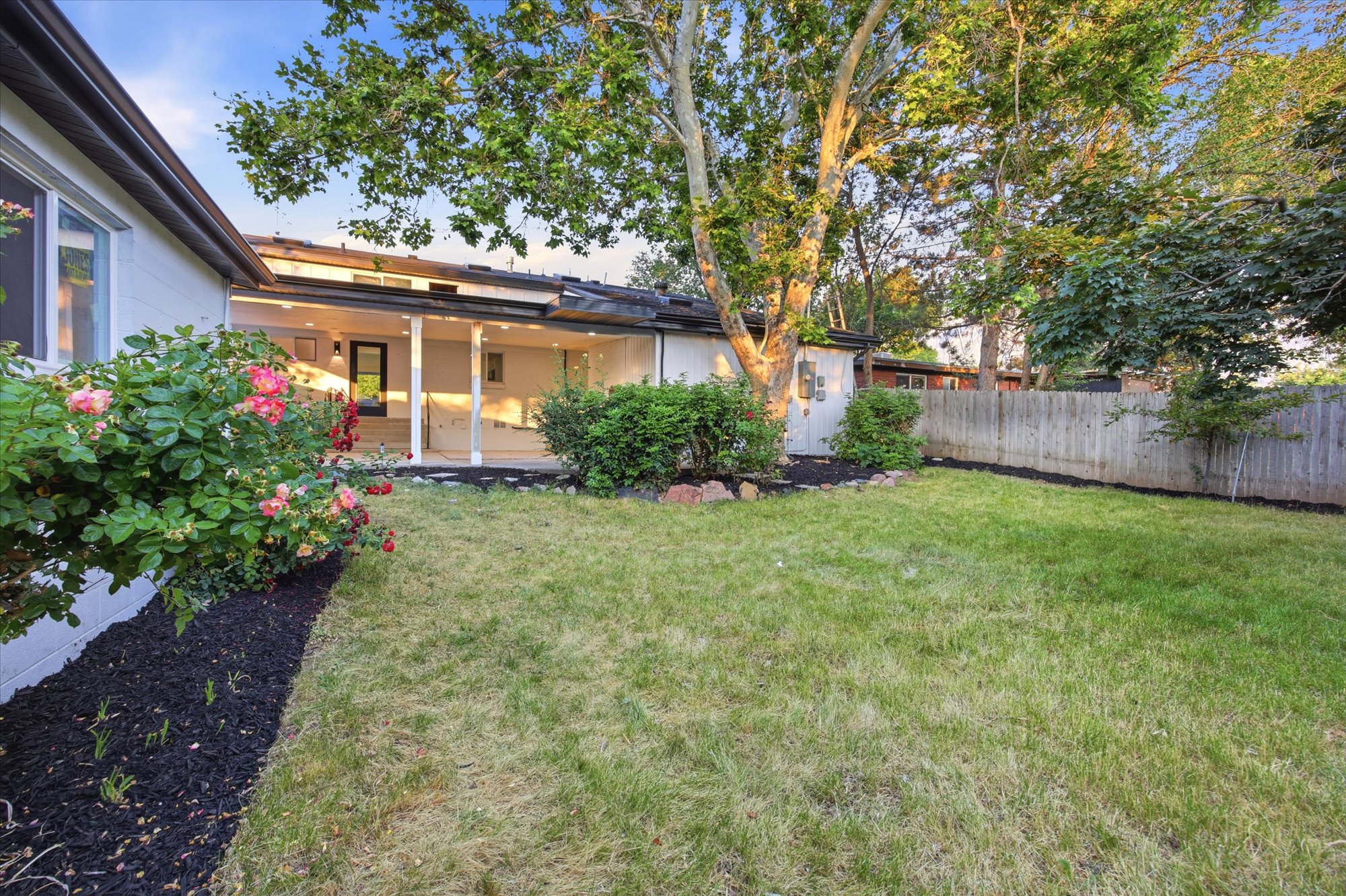 Expansive rear yard featuring a covered patio, privacy fencing, mature trees, and landscaping with flowering shrubs