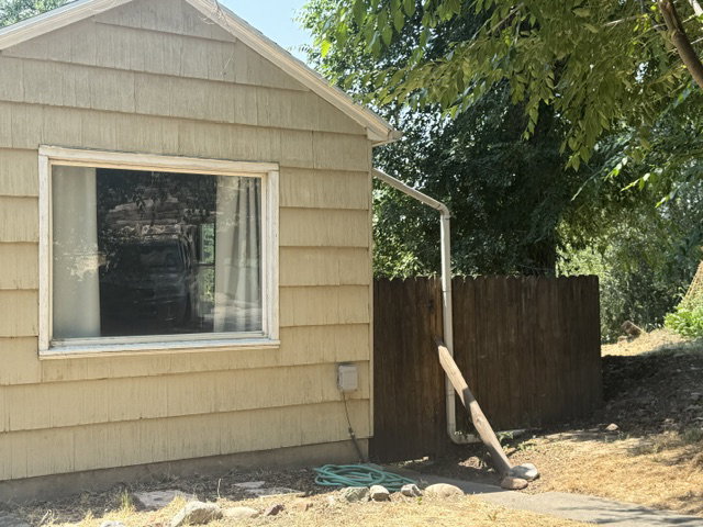Beige shingle siding exterior featuring a rectangular window with white trim, a brown wood fence, and mature trees in the background
