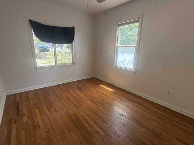 Hardwood flooring throughout, featuring two windows, white walls, and a ceiling fan