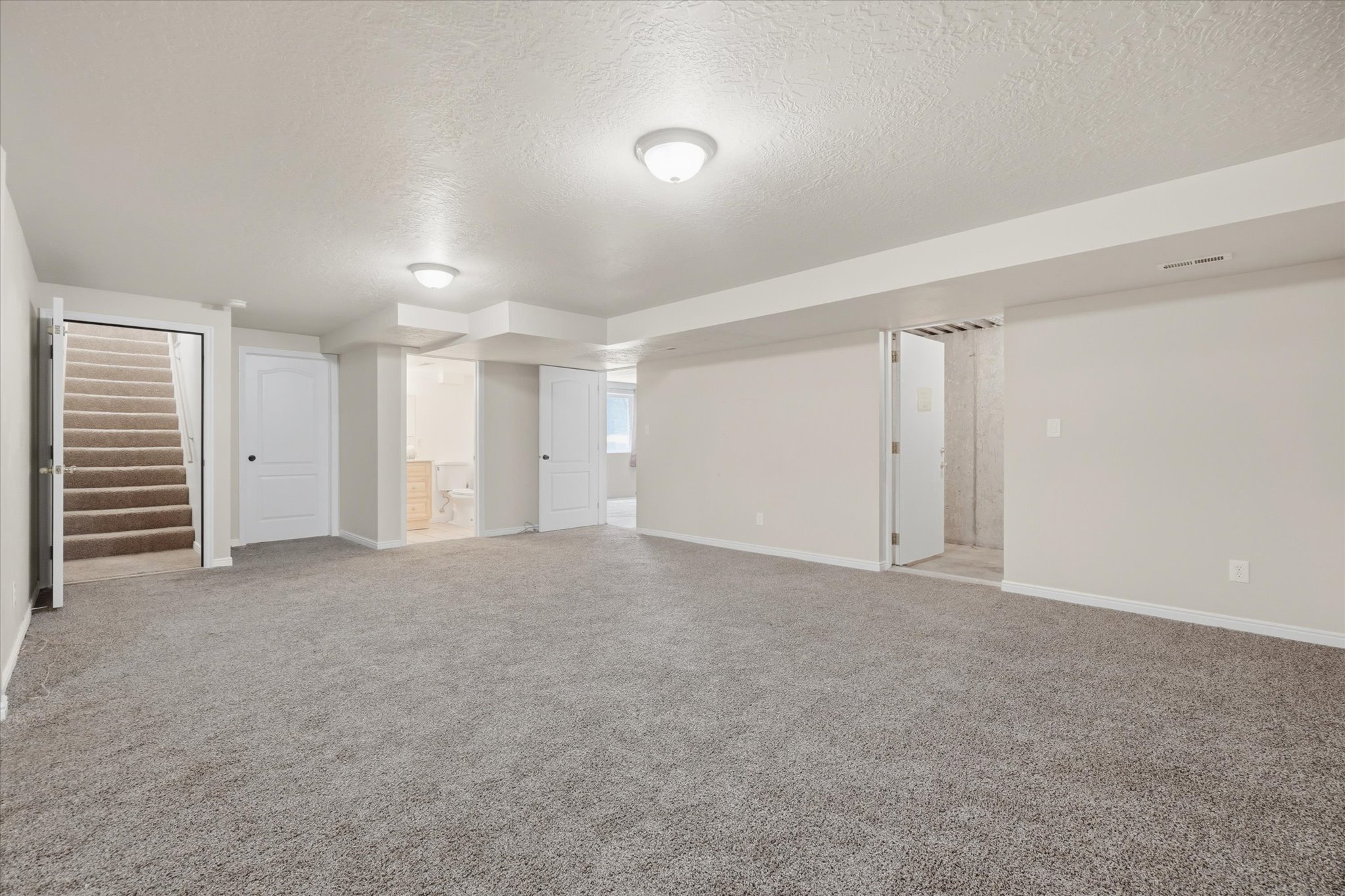 Spacious carpeted room featuring recessed lighting, a fully finished staircase, multiple white panel doors, and a partial bathroom with a built-in vanity